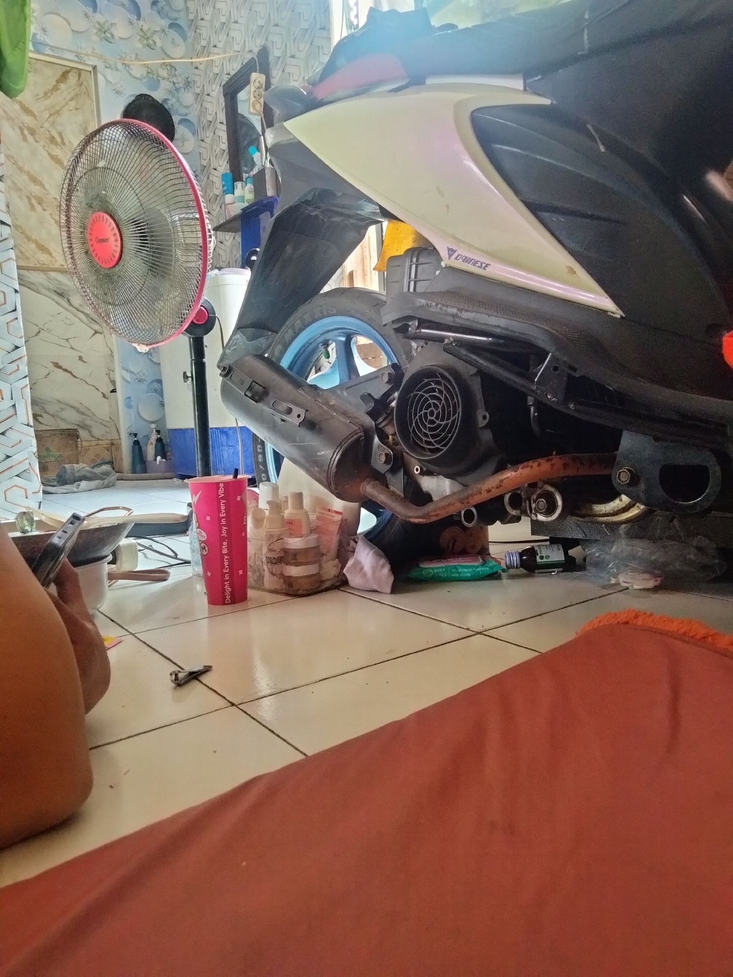 The image shows the interior of a room, likely a home, in Cariu, Indonesia. A motorcycle is parked in the center of the frame, with its engine and exhaust visible. To the left, a floor fan with a red trim is positioned near a patterned wall. In the foreground, a person's legs are visible, and a pink cup and various bottles are scattered on the tiled floor. A nail clipper is also on the floor near the person. The overall environment appears cluttered and suggests a casual, lived-in space.