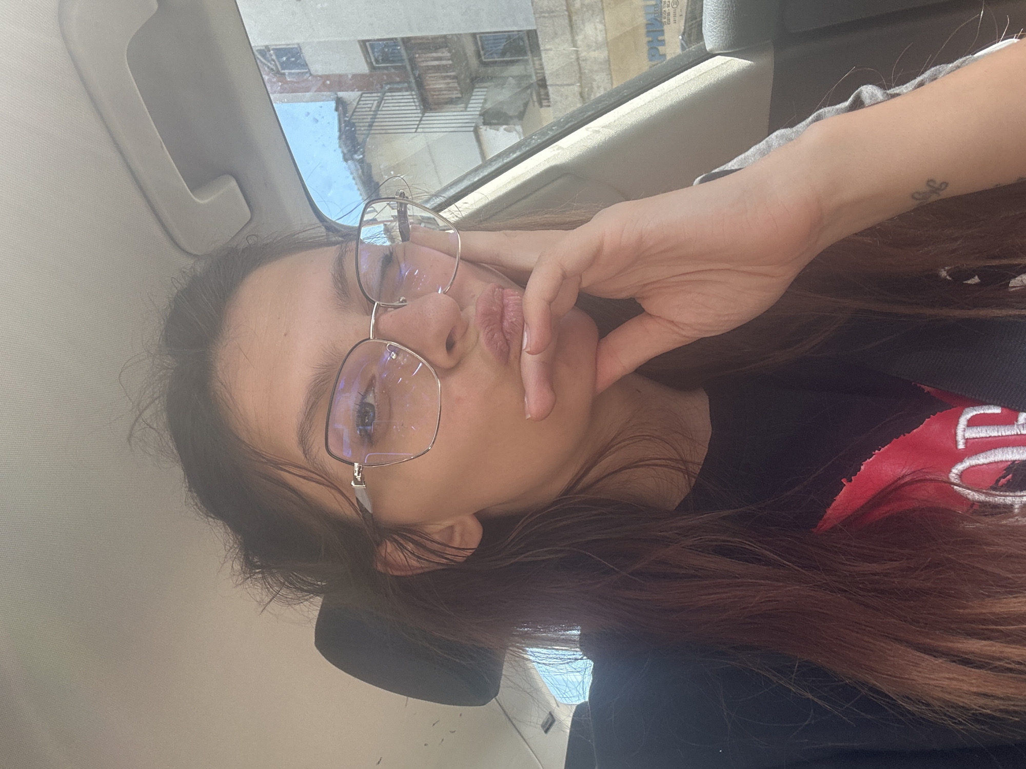 The image shows a young woman in the passenger seat of a car, with buildings visible through the window. She is wearing glasses and a black t-shirt with a red design. Her hand is resting on her chin, and she is looking towards the camera with her lips pursed. The lighting suggests it is daytime. The location is identified as Tryavna, Bulgaria.