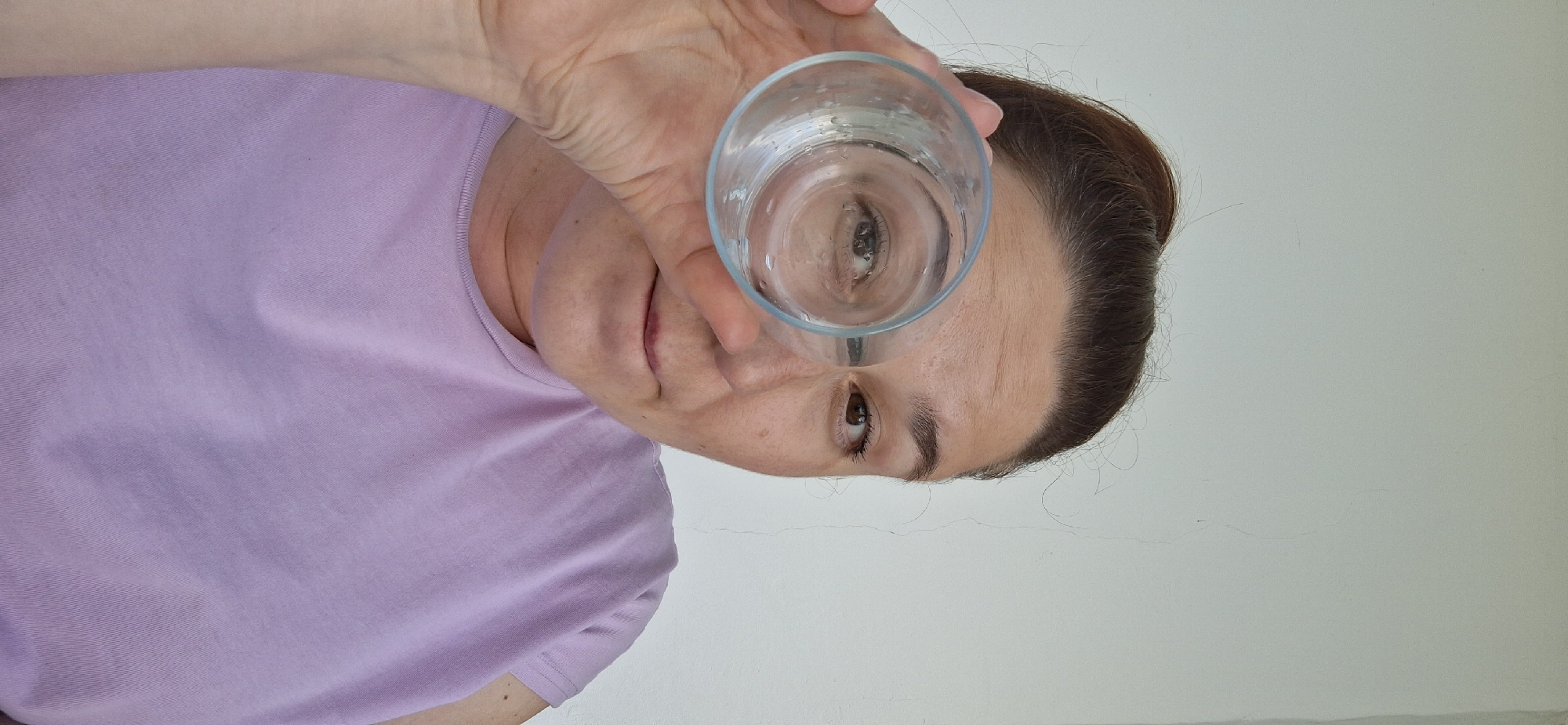 A person is holding a glass of water in front of their face, with their eye visible through the water. The person is wearing a purple t-shirt. The background is a plain white wall. The image was taken from a low angle. There is no evidence of Chaikovsky, Russia in the image.
