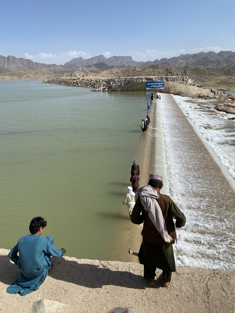 The image shows a group of people walking across a dam or weir in Kim, Afghanistan. The structure creates a waterfall on one side. Mountains are visible in the background under a clear sky. One man sits on the edge of the structure in the foreground while another walks along the dam, his back to the viewer. Others are walking on the dam away from the foreground. A sign in Arabic is visible at the end of the structure.