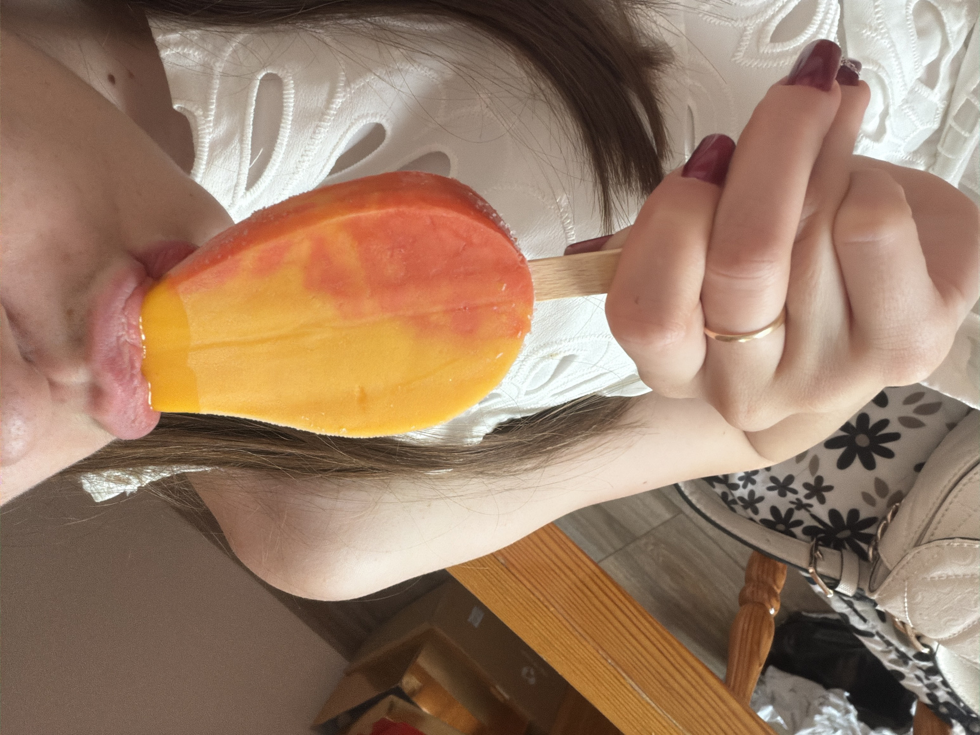 An individual is depicted indoors, consuming a bi-colored popsicle. The upper portion of the person's head, neck, shoulder, and arm are visible. The individual's lips are parted, making contact with the yellow and orange popsicle. The right hand, with dark red manicured nails and a plain gold ring on the ring finger, holds the popsicle stick. Long, dark brown hair extends over the visible shoulder. The individual wears a white garment with a decorative cut-out pattern on the fabric.
The background includes a light-colored wall or ceiling in the upper left. A light brown wooden chair or bench with horizontal slats is positioned in the lower left. Beneath this, a segment of greyish-brown flooring, a light-colored handbag, and a floral-patterned fabric are discernible. There are no visual elements within the image to confirm the location as the city of Varna, Bulgaria.