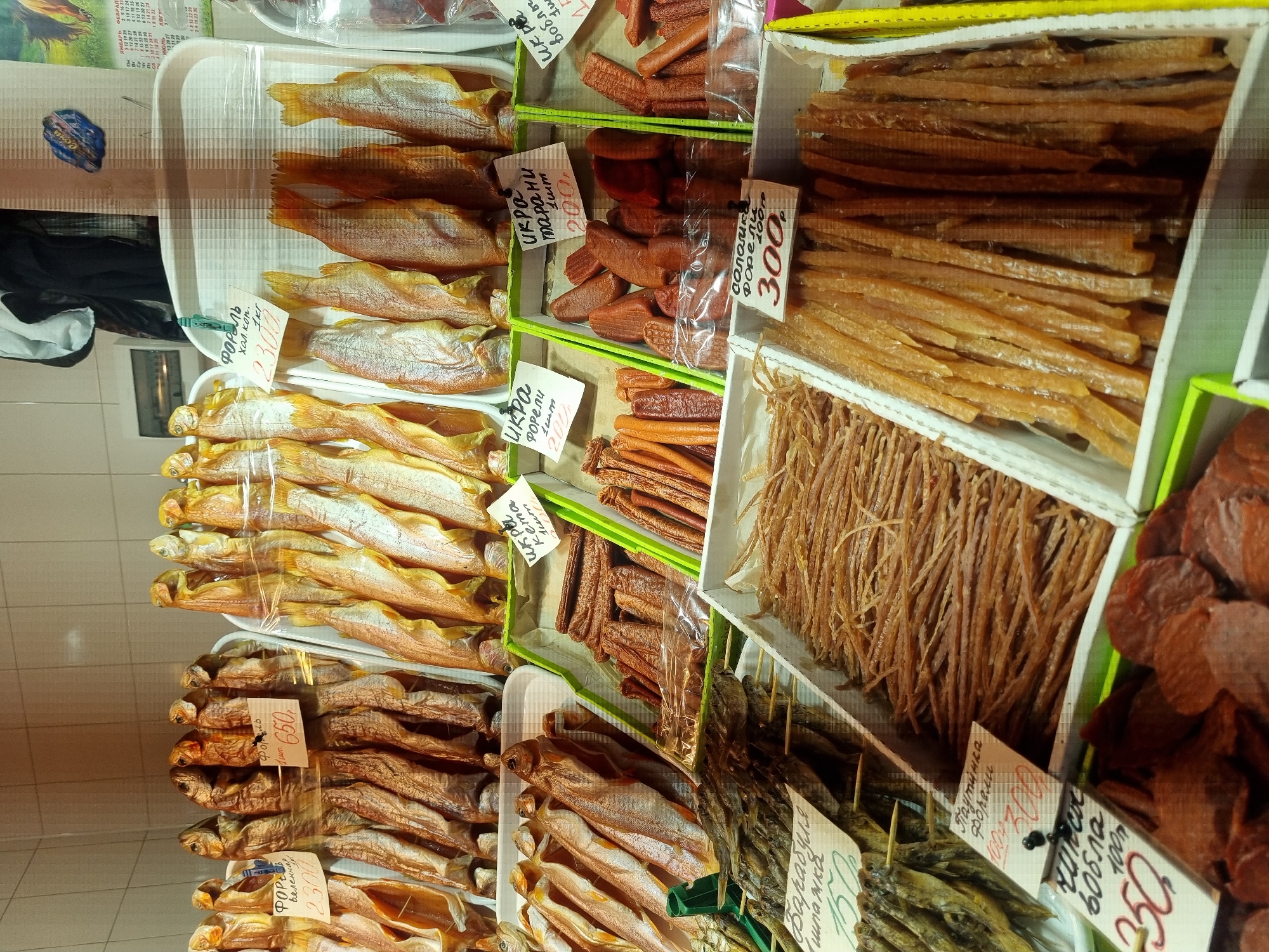 The image shows a display of various dried and smoked fish and seafood products in a retail setting. The location is likely Sochi, Russia, based on the Russian script on the price tags and product names.
The display includes different types of fish (e.g., trout, roach) presented in trays and boxes. Some items are labeled with price tags indicating the cost per kilogram or piece. The fish range in color from golden to dark brown, indicative of different curing and smoking processes. Besides whole fish, there are also dried fish sticks, and circular pieces of some processed seafood. Some of the products are packaged in clear plastic.
The overall environment appears to be a market or a store specializing in seafood or smoked products. The background has tiled walls. The image suggests a variety of options for customers looking to purchase dried or smoked fish products.