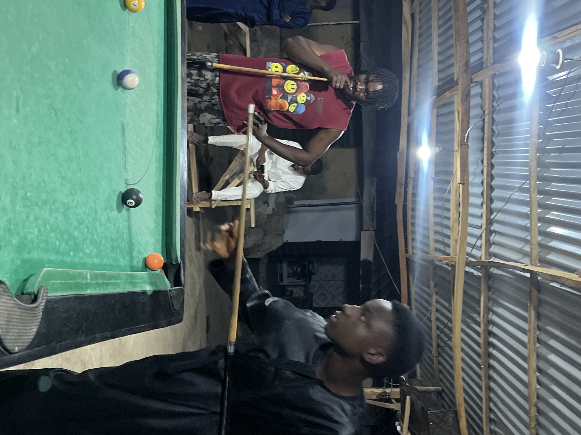 This vertical, indoor photograph captures several men engaged in a game of pool under artificial lighting, likely at night or in a dimly lit interior. The setting appears casual, possibly an informal entertainment spot, featuring corrugated metal walls supported by wooden framing.
In the foreground, a young man in a dark, long-sleeved top stands with a pool cue, looking towards the table with a focused expression. To his right, another man in a red t-shirt adorned with various smiley face graphics (yellow, orange, blue) leans over the green-felted pool table, holding a cue as if preparing or executing a shot. He has dreadlocks.
On the pool table, several balls are visible, including a yellow solid ball, a white and purple striped ball, a black 8-ball, and an orange solid ball. The table has black bumpers.
In the background, two other men are present. One, dressed in a dark blue outfit, stands with his arms crossed, observing the game. Another man in a white outfit is seated on a simple wooden stool or structure, also watching.
Multiple bare light bulbs emit bright glares, illuminating the scene and casting strong shadows, highlighting the artificial nature of the light source. The overall ambiance is one of relaxed social interaction and concentration on the game. No legible text is present beyond the smiley face graphics on the red shirt. The location cues suggest a functional, unadorned space.