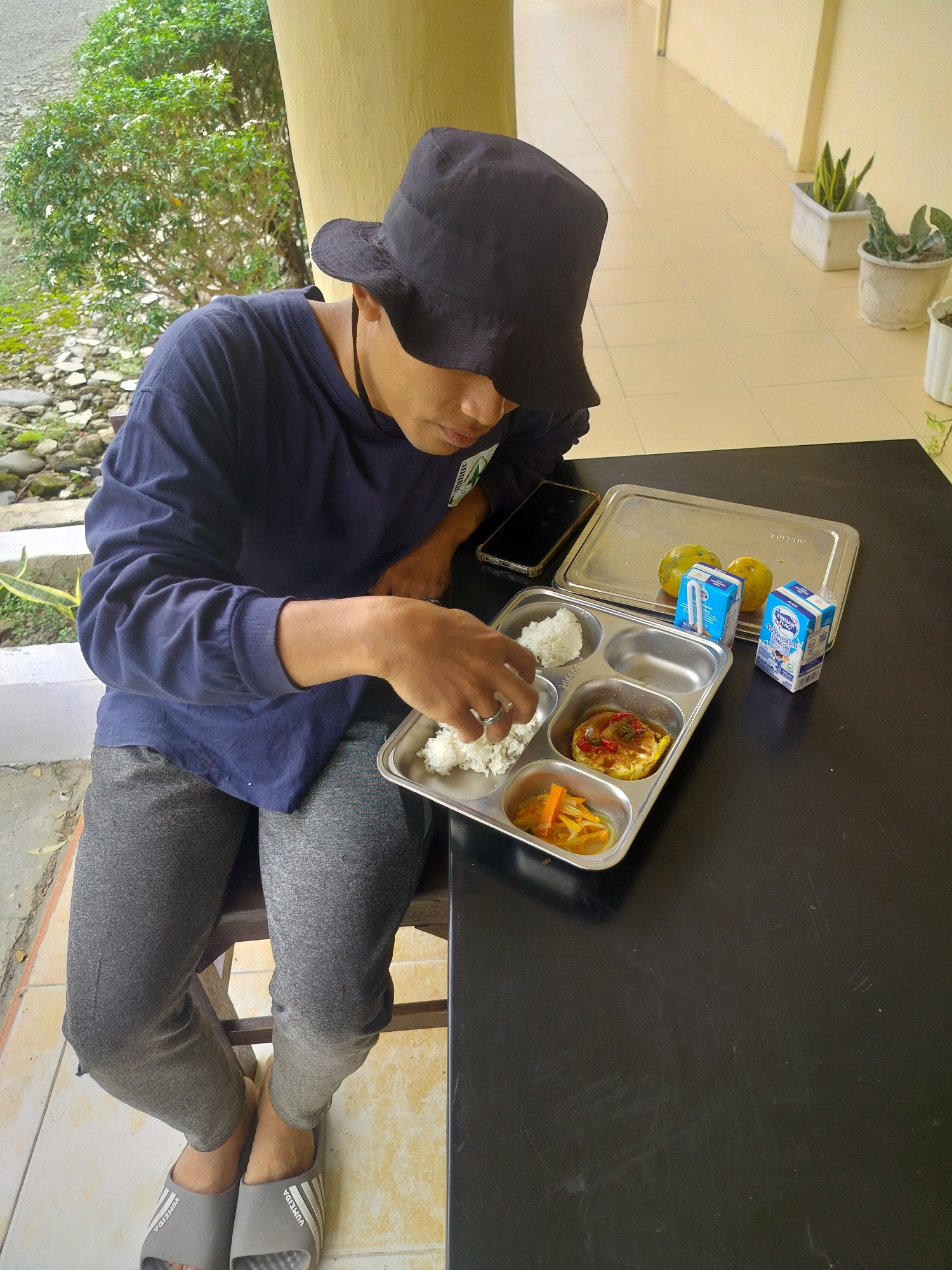 A young person wearing a black bucket hat and a navy blue long-sleeved shirt sits at a black table with a meal tray. The meal tray contains rice, an egg dish with sauce, and pickled vegetables. Next to the tray are two small cartons of milk and two oranges on a larger metal tray. A smartphone is also on the table. The person is reaching into the rice compartment of the meal tray with their right hand. The background shows an outdoor area with a yellow building and potted plants, suggesting a location in Lawebekung, Indonesia. The person is wearing grey sweatpants and grey slippers.