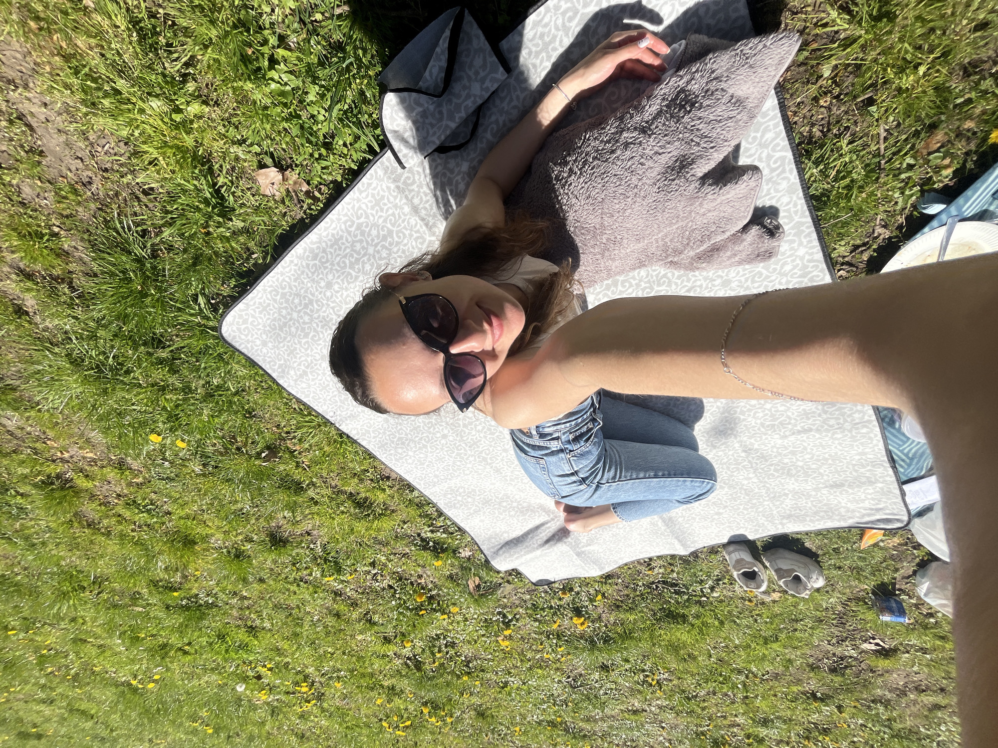 A woman wearing jeans and sunglasses is lying on a patterned blanket in a grassy area, possibly a park. The woman is facing the camera, and the perspective suggests a selfie. A grey blanket or towel is next to her, and there are shoes near the edge of the blanket. Additional items and bags are nearby. The location is likely a park in Saarbrücken, Germany, as instructed, but specific landmarks are not visible in the image.