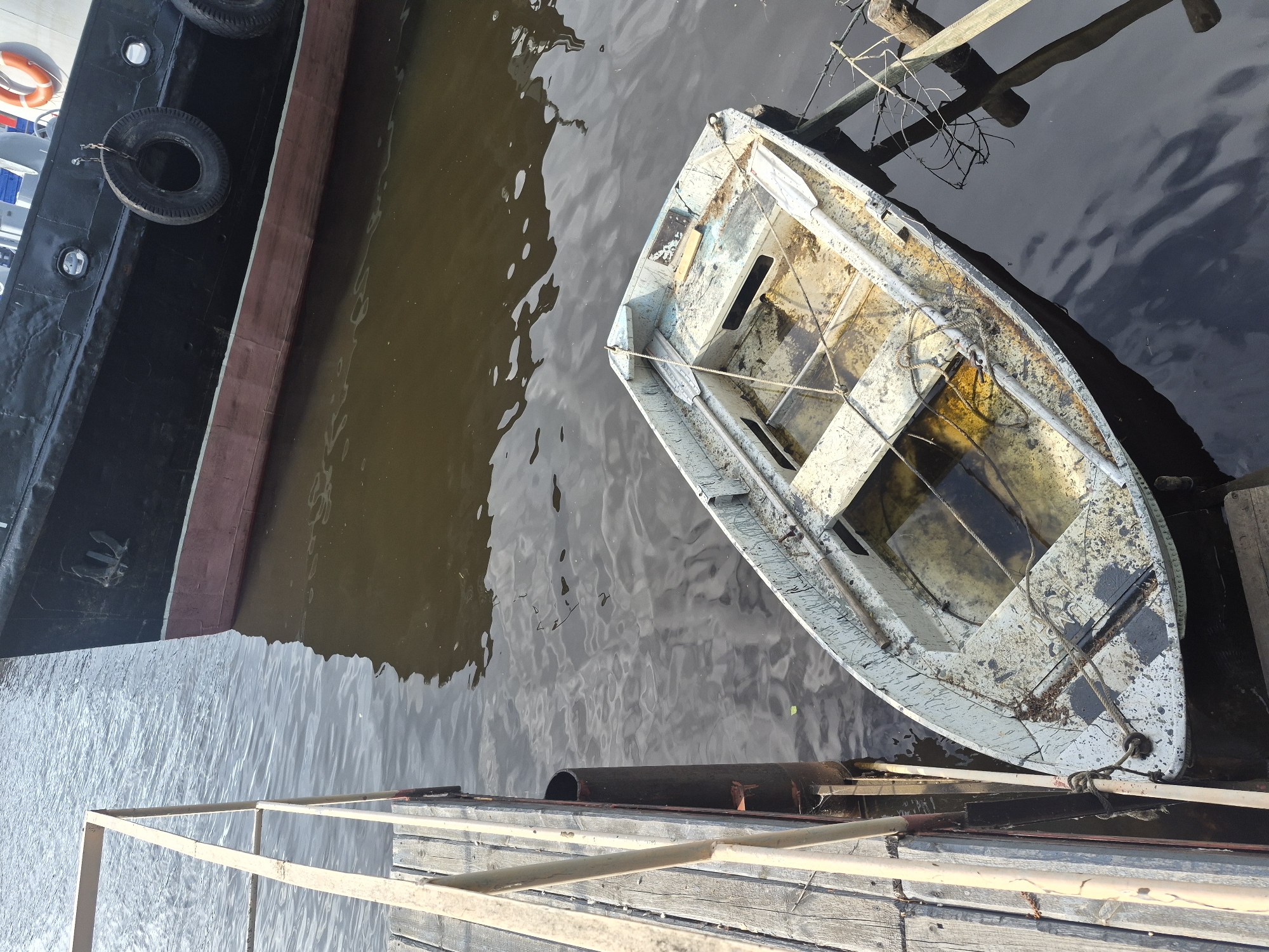 The image depicts a small, old, light-colored boat partially filled with water docked next to a larger black ship. The small boat is tied to a wooden dock. The location appears to be Chaikovsky, Russia, based on visual details. The water surrounding the boats is murky. The dock has a simple railing. There are no people visible in the image.