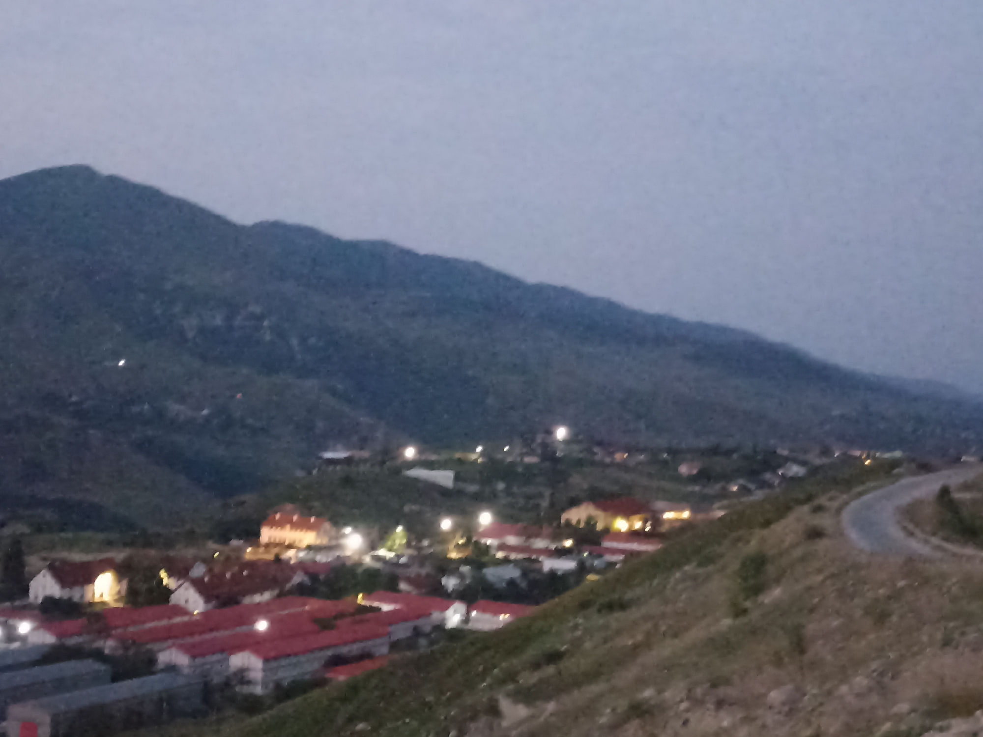 The image shows an elevated view of a town or village at dusk, likely in the city of Kuzun, Azerbaijan, given the context. The foreground is dominated by a steep, grassy hillside with scattered rocks and a winding paved road that curves out of view to the right. In the middle ground and background, a cluster of buildings with red tile roofs is visible, illuminated by artificial lights. Some buildings appear to be residential, with multiple stories and illuminated windows. Other structures, possibly industrial or agricultural, are also present, some with dark, elongated roofs. The town is nestled in a valley, with rolling, sparsely vegetated hills and mountains forming the backdrop. The sky is a pale, overcast grey, characteristic of twilight. No people are visible in the image. The overall impression is one of a rural or semi-rural settlement in the early evening.