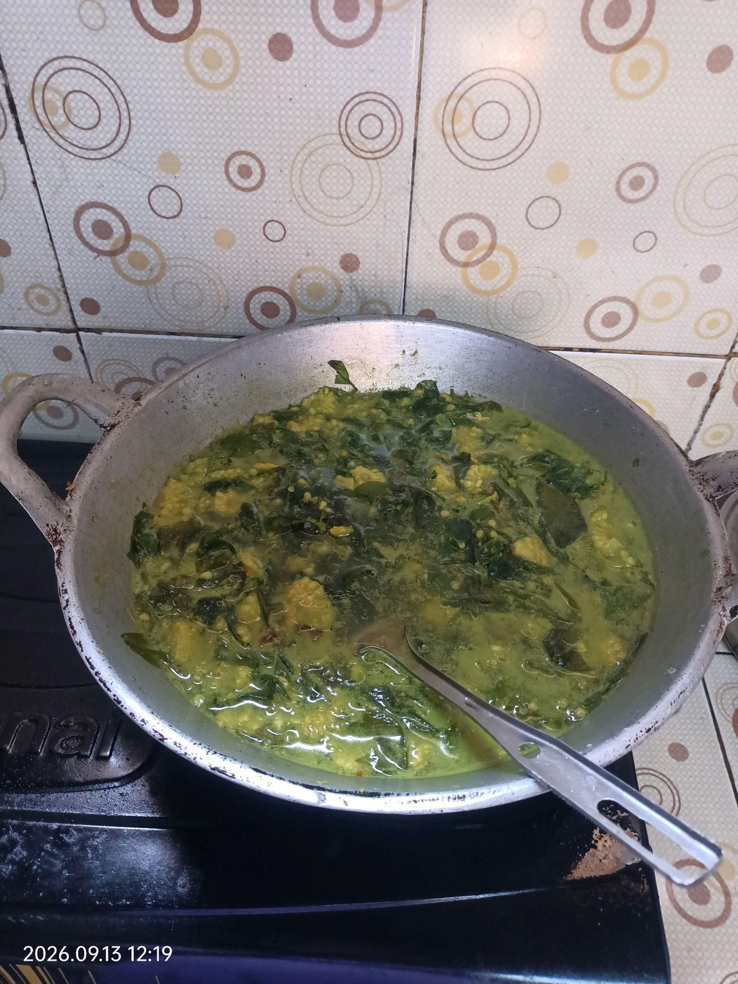 A high-angle, close-up shot captures a traditional Indonesian dish being cooked in a silver metal wok on a black stovetop. The wok is filled with a vibrant green curry, containing leafy vegetables and pieces of what appear to be tofu or chicken. A silver spoon rests inside the wok, with its handle extending out. The background features a tiled wall with a circular, abstract pattern in shades of beige, brown, and cream. The image is dated "2026.09.13 12:19".