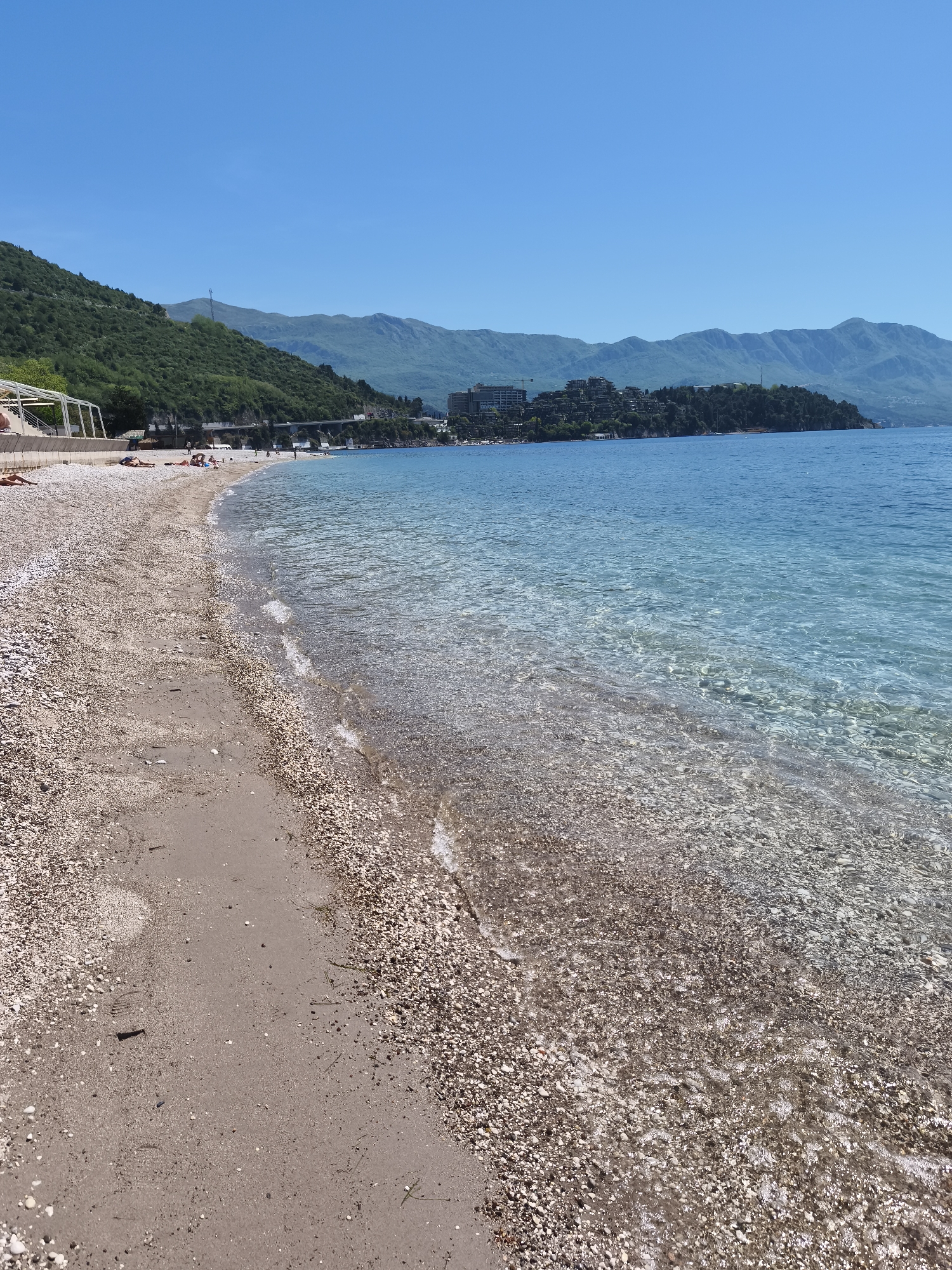 The image captures a picturesque coastal scene on a sunny day in Budva, Montenegro. In the foreground, a tranquil beach curves gently, composed primarily of light-colored pebbles mixed with patches of sand. The clear, shallow water, transitioning from turquoise to a deeper blue, reveals the pebbly seabed beneath the gentle waves.
Several people are visible on the beach, engaging in leisure activities such as sunbathing on towels or walking along the water's edge. To the far left, a white, open-framed structure, possibly a shaded area or part of a beach bar, stands near the back of the beach.
Beyond the shoreline, lush green, forested hills rise steeply, with a road winding along their base. In the mid-ground, a headland covered with buildings, likely part of a town or resort, extends into the sea. Majestic mountains form a distant backdrop, stretching across the horizon under a vast, cloudless blue sky. The bright, clear sunlight indicates it is daytime, contributing to the serene and inviting atmosphere of the beach. No visible text is present in the image.