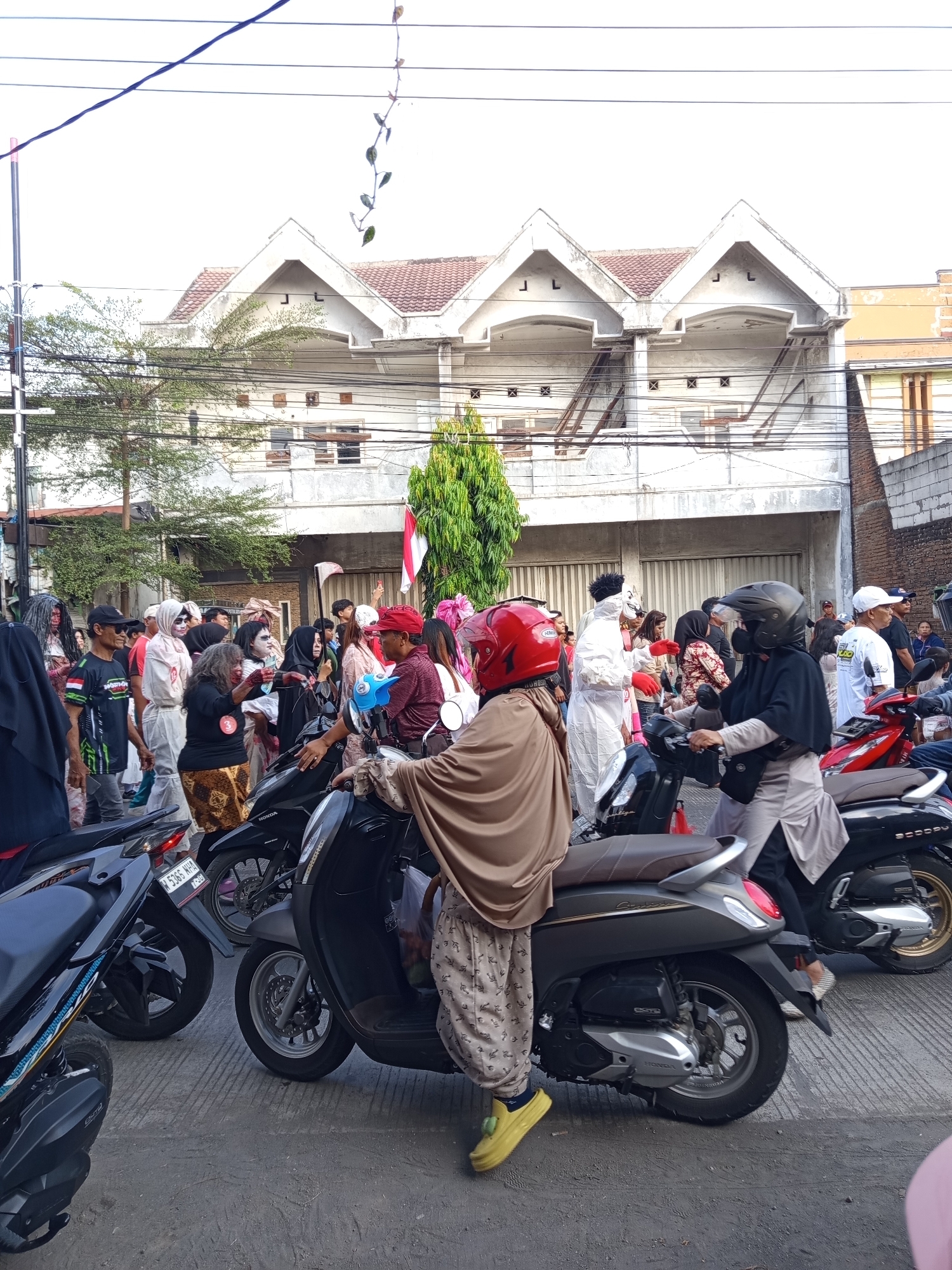 The image depicts a street scene in Suko, Indonesia, with a crowd of people gathered. Many individuals are dressed in costumes, suggesting a celebration or event. Several people are riding or standing near motorcycles. In the background, a two-story building with a distinctive architectural style, featuring multiple gable roofs, is visible. The sky is bright, indicating daytime. The overall atmosphere appears to be festive and social.