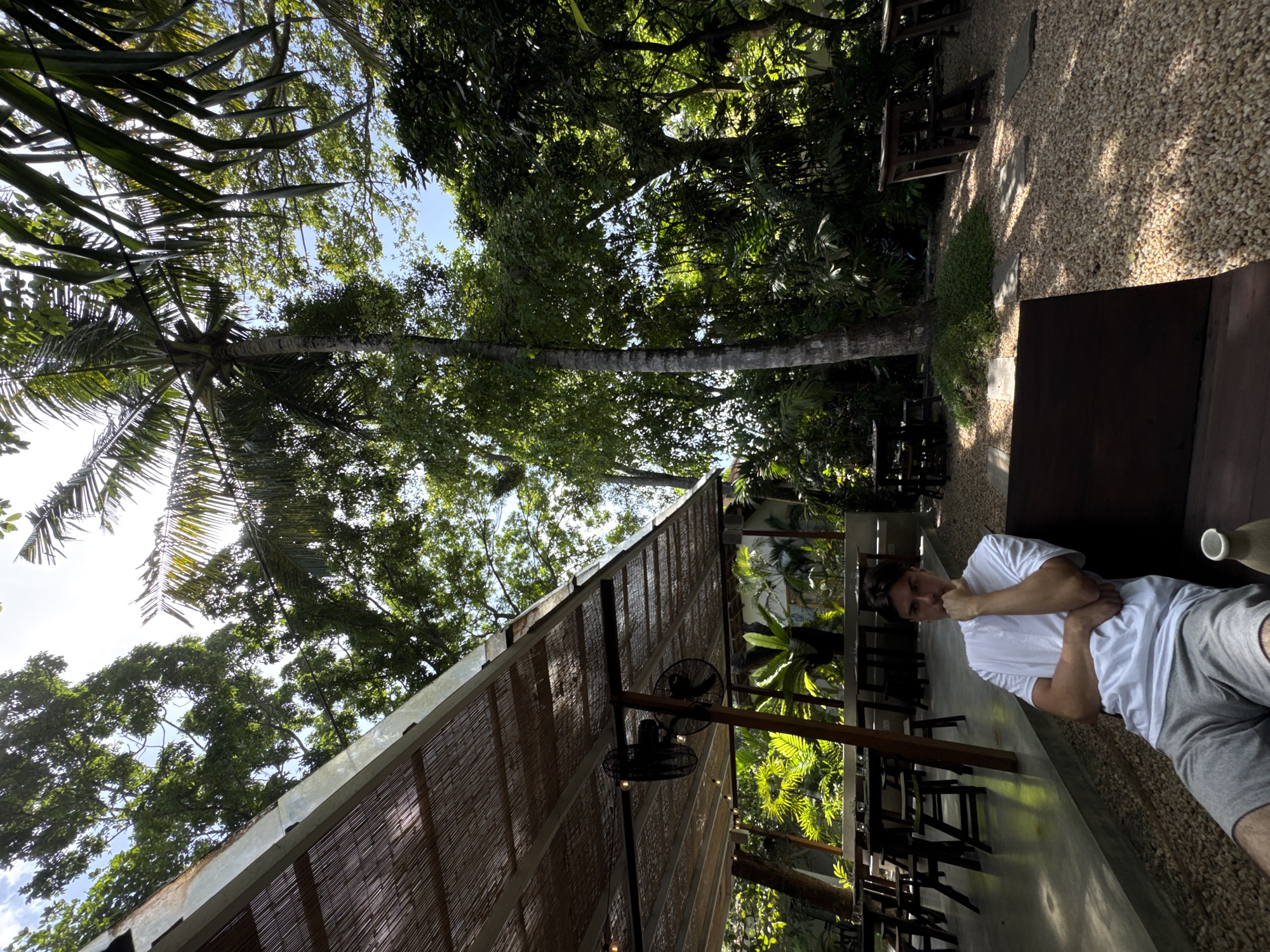 A young adult male, appearing to be in his late teens or early twenties, is seated on a dark wooden bench in an outdoor cafe. He is wearing a white t-shirt and grey shorts, with his right arm resting on his lap and his left elbow on the bench, his chin in his hand. The cafe has a thatched roof supported by metal beams, with two industrial fans mounted underneath. Tables and chairs are visible in the background, some set for dining. The ground is covered with light-colored pebbles. The surrounding environment is lush and green, with tall trees, including a prominent palm tree, and dense foliage, suggesting a tropical location. The bright sunlight filtering through the leaves creates dappled shadows. The scene is set in the city of Ahangama, Sri Lanka.