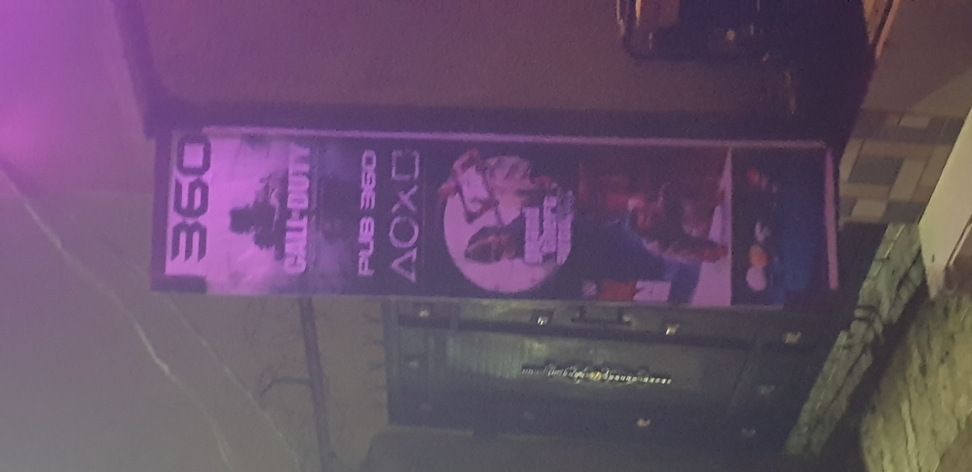 The image shows a vertical sign with gaming-related graphics and text, illuminated by purple lighting. The sign features the number "360" at the top, followed by an image of a silhouetted figure and the text "CALL-DUTY". Below this is "PUB 360" and the PlayStation logo. Further down, there is a circular graphic that appears to be from Grand Theft Auto, and another image of a figure. The sign is positioned outdoors, with a dark doorway or gate visible to the left and a building structure on the right. The scene appears to be at street level in the city of Narayi, Nigeria, possibly indicating a gaming center or pub.