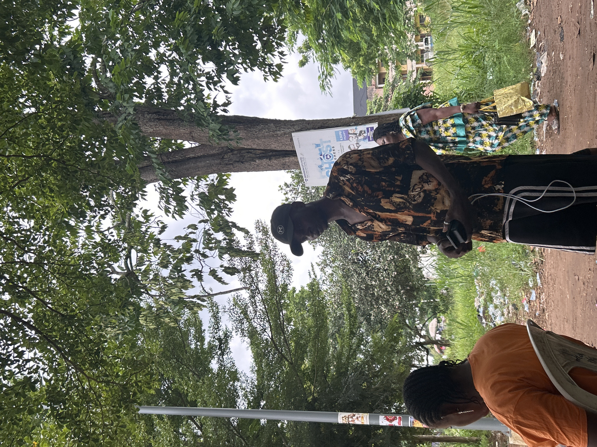 In Ihiagwa, Nigeria, a scene unfolds outdoors with a focus on individuals against a backdrop of trees and urban elements. The image features a man wearing a dark baseball cap and a patterned short-sleeved shirt. He is holding a small black object in his right hand and appears to be in conversation or engaged with his surroundings. To his left, a woman is seated, facing away from the camera, wearing an orange t-shirt. Further to the right, a woman dressed in a colorful, patterned outfit with a turquoise trim is visible. She is carrying a gold-colored bag and a black handbag. In the background, tall green grass and some debris are scattered on the ground. A pole with stickers is present on the left, and a large tree with a sign attached to its trunk stands prominently. Buildings and a vehicle are faintly visible in the distance, suggesting a populated area within the city. The overall environment suggests a moderately overcast day, with lush green foliage dominating the upper portion of the frame.