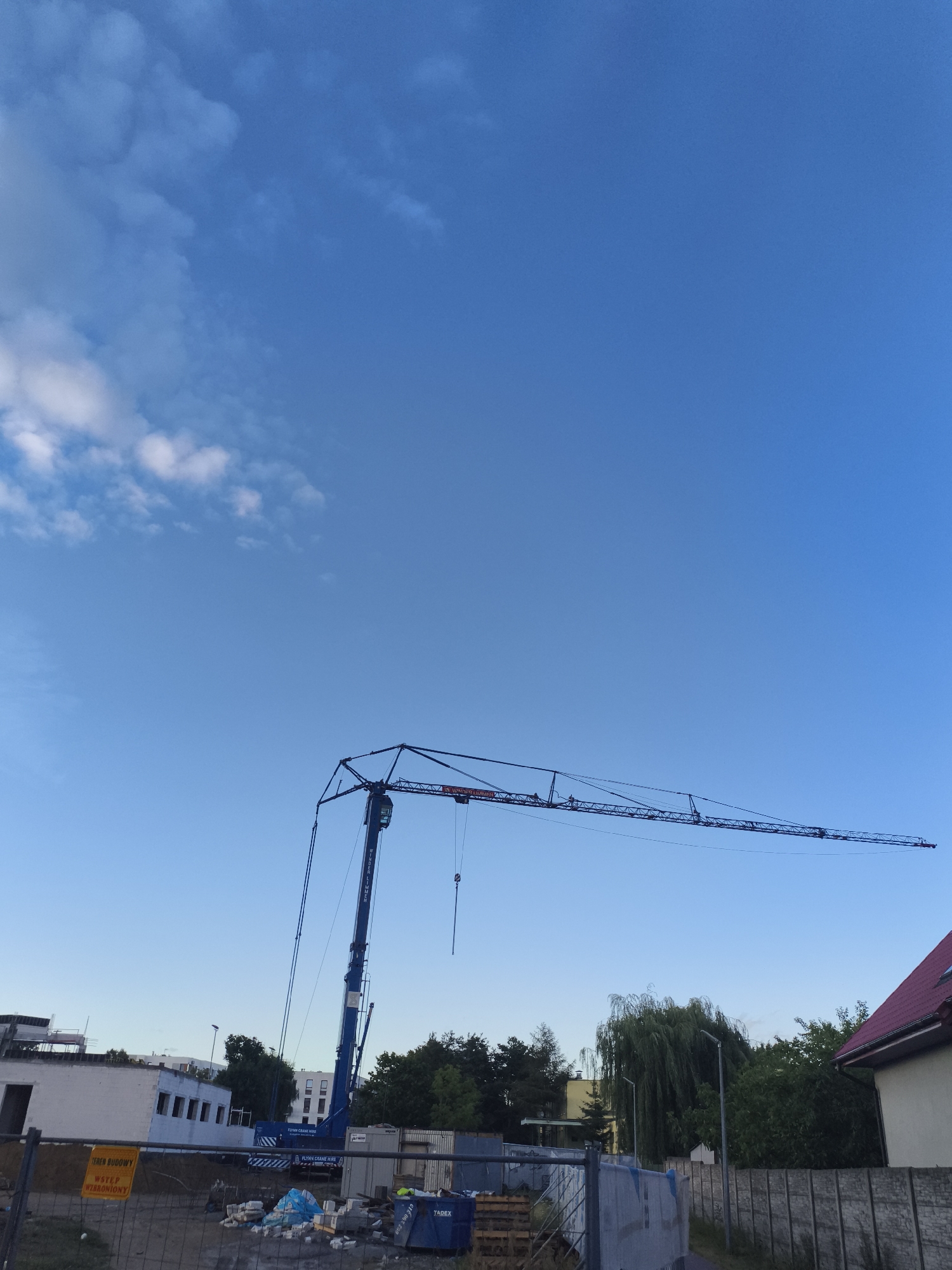 This is an image of a construction site with a large blue crane dominating the center of the frame. The crane's arm extends horizontally across the upper portion of the image against a clear blue sky with a few scattered white clouds. In the foreground, there is a chain-link fence surrounding the site, with debris and construction materials visible behind it. A yellow sign with Polish text that reads "TEREN BUDOWY WSTĘP WZBRONIONY" (construction site, entry forbidden) is attached to the fence. To the left, there is a partially constructed building with exposed white walls and windows. Trees and other buildings are visible in the background, suggesting an urban or suburban environment. The overall scene depicts an active construction area in Nowy Dwór Mazowiecki, Poland, under a bright sky.