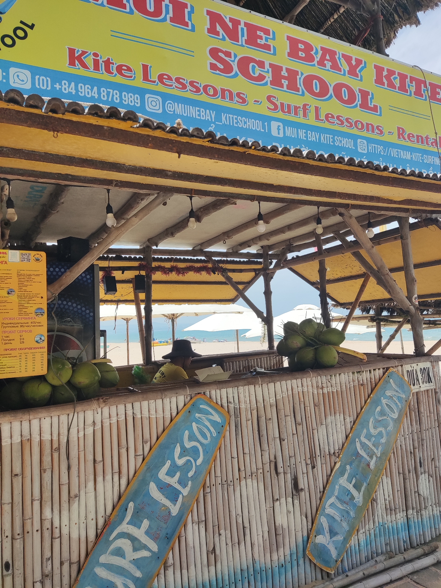 The image depicts an outdoor scene at a beach establishment, likely a surf school and rental shop. A large yellow banner with red and blue lettering across the top announces "MUI NE BAY KITE SCHOOL" and lists "Kite Lessons - Surf Lessons - Rental". Below this banner, a bamboo-constructed counter is visible, displaying a bunch of green coconuts. Behind the counter, a person wearing a yellow shirt and a dark hat is seated, facing away from the camera. Several beach umbrellas and lounge chairs are set up on the sandy beach in the background, with the ocean and a clear sky beyond. Two blue and yellow wooden signs with white lettering, reading "SURF LESSON" and "SURF LESSON", are leaning against the bamboo wall on the right side of the frame. The overall atmosphere suggests a tropical, tourist-oriented beach setting.