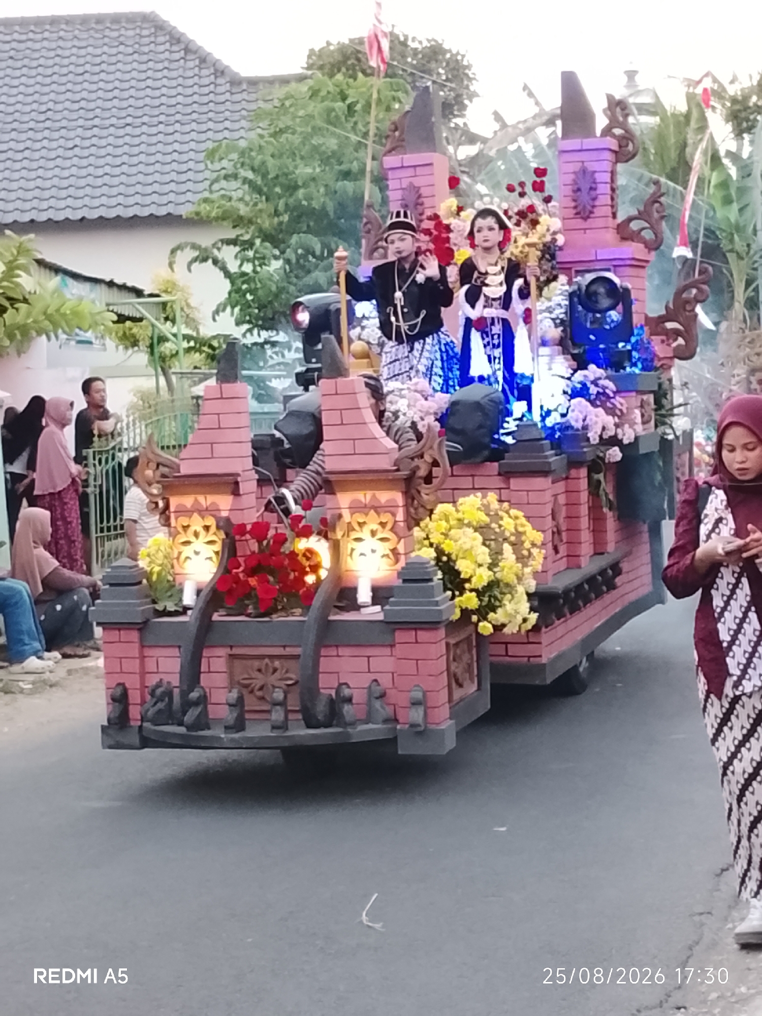 This image captures a parade float decorated with traditional Indonesian attire and flowers. Two individuals, a male and a female, are dressed in formal Javanese wedding attire and are standing atop the float. The float itself is designed to resemble a brick structure with decorative elements and illuminated with blue and pink lights. In the background, several people are gathered, some observing the parade and others participating. The scene appears to be taking place in a residential area, with houses and trees visible. The timestamp "25/08/2026 17:30" suggests this event occurred on August 25, 2026, at 5:30 PM. The location is identified as Semanding, Indonesia.