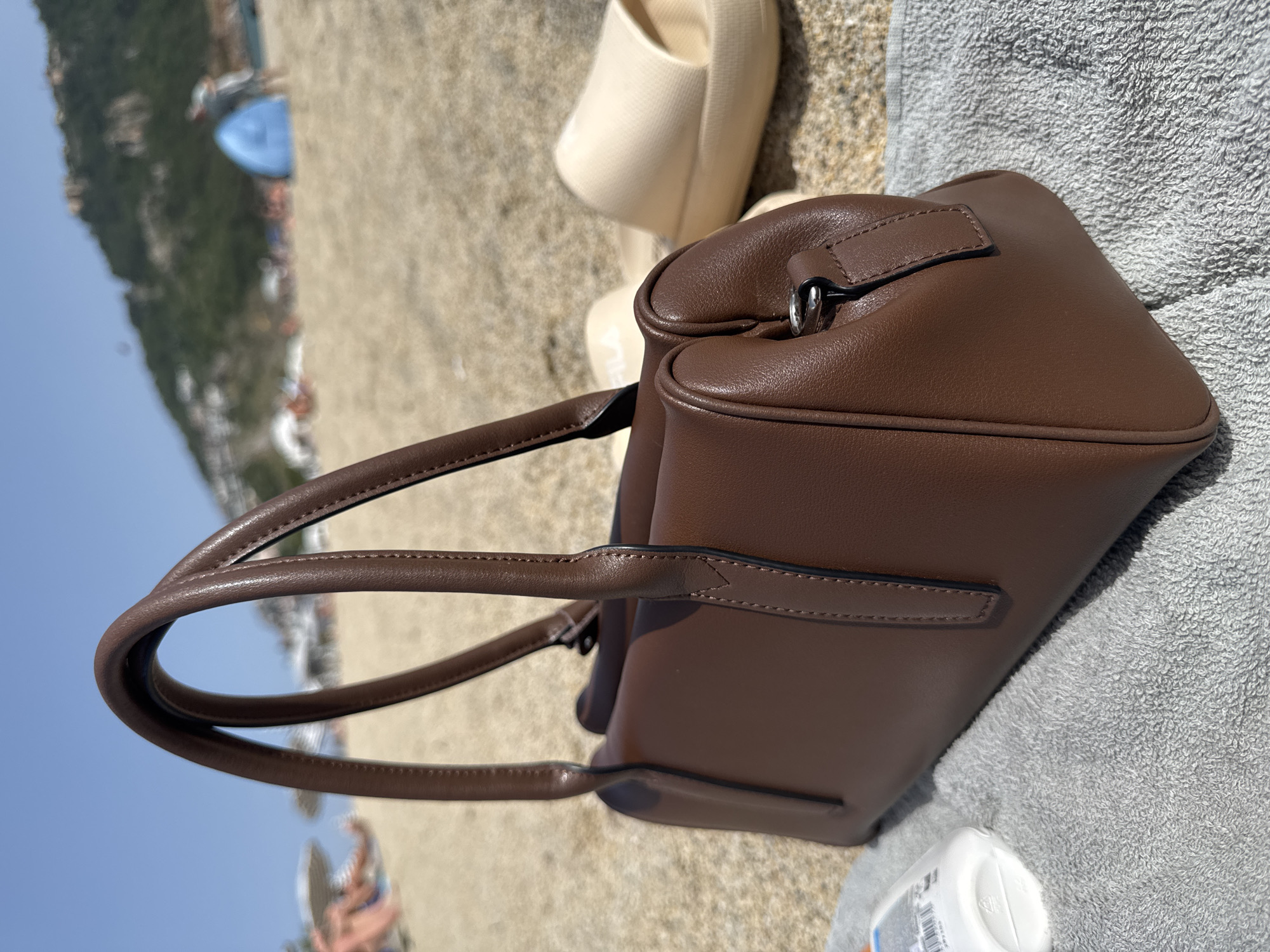 On a sunny day at a beach in Alanya, Turkey, a brown leather handbag rests on a gray towel next to beige sandals and a white bottle. The beach stretches toward the Mediterranean Sea, visible under a clear blue sky. A distant mountainside rises behind the beach, and blurry figures of people are visible in the background.