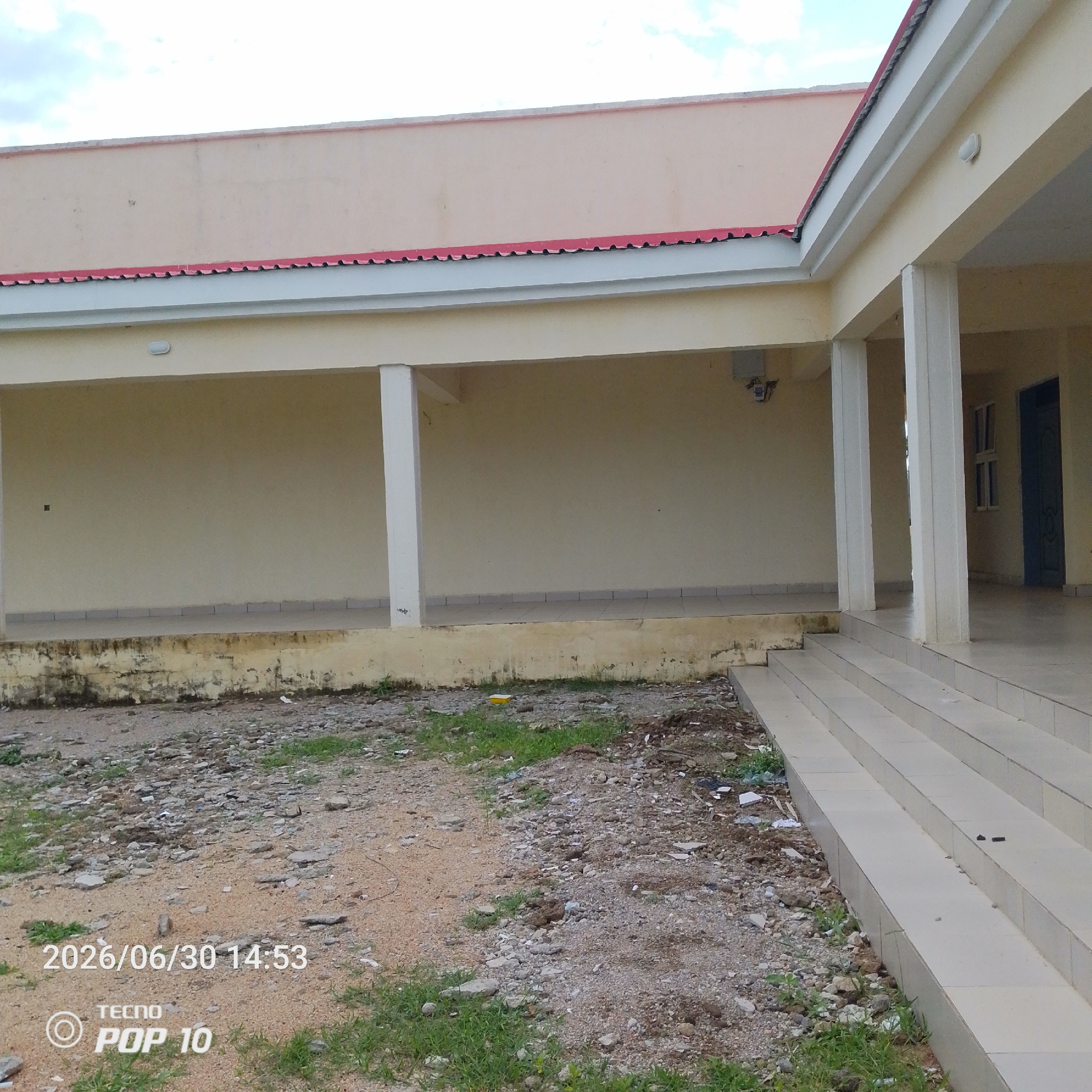 This image depicts an outdoor scene in Jalingo, Nigeria, likely on a bright day with some clouds visible in the sky. The primary subject is a building with a pale yellow exterior wall and a light-colored roof with a red trim. The building features a covered walkway supported by white columns, creating a shaded area.
In front of the building, there is a ground surface composed of gravel, sand, and scattered debris, with some patches of green grass growing. A tiled staircase leads up to the building's entrance. The overall impression is that of a somewhat unkempt but functional building, possibly a school or community center, with evidence of recent construction or disuse in the surrounding area.
There are no people visible in the image. Visible objects include the building itself, the staircase, columns, an outdoor light fixture on the wall, a small electrical box, and a dark blue door with a window in the distance. The ground is covered with various sizes of stones and dirt.
The visible text "2026/06/30 14:53" indicates the date and time the photo was taken. Below this, "TECNO POP 10" suggests the brand and model of the phone used to capture the image. The scene appears to be from the afternoon, given the time stamp. The weather seems fair, with the sky suggesting it is not raining. The location context of Jalingo, Nigeria, is provided, adding a geographical layer to the scene.