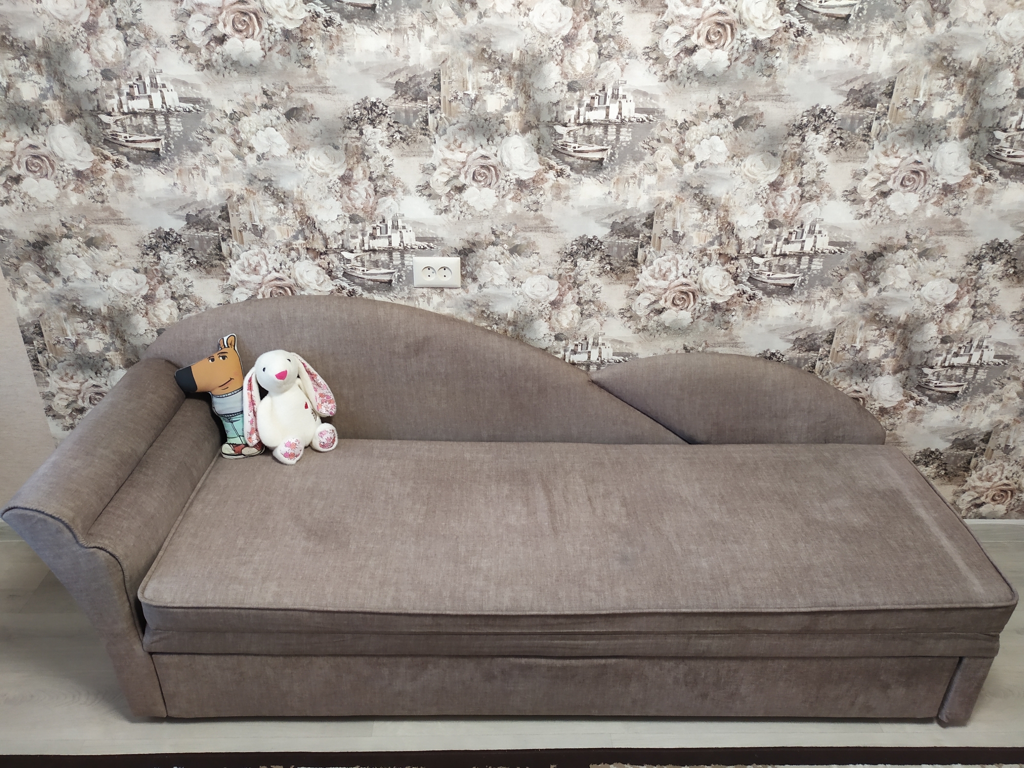 A brown chaise lounge sits in front of a floral and landscape-themed wallpaper. Two plush toys, a dog and a white rabbit, are placed on the left side of the chaise lounge. The chaise lounge has a slightly curved backrest and is upholstered in a textured brown fabric. The wallpaper features a pattern of roses and a coastal landscape with buildings and boats. Two white electrical outlets are visible on the wallpaper. The flooring beneath the chaise lounge appears to be light-colored laminate or wood.