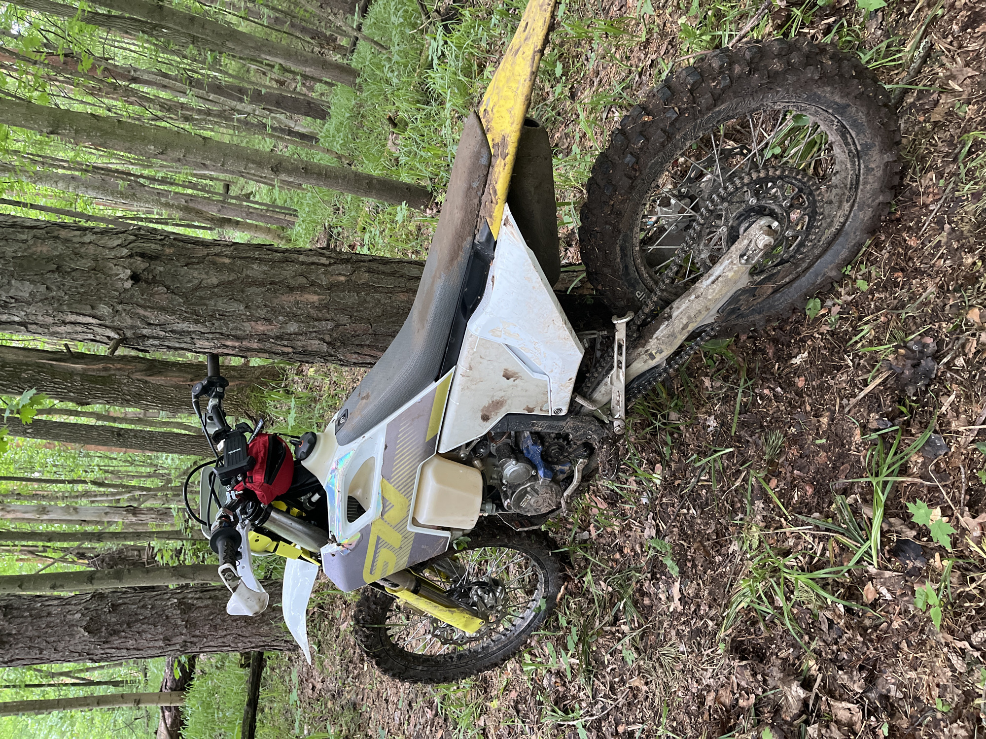 The image shows a dirt bike lying on its side in a wooded area. The bike is white and yellow, and appears to be a Husqvarna. The front wheel is turned slightly towards the left. The tires are muddy. A red pouch is attached to the handlebars. The background consists of trees and undergrowth. The location is not identifiable as Gruzinskii, Russia, from the image.