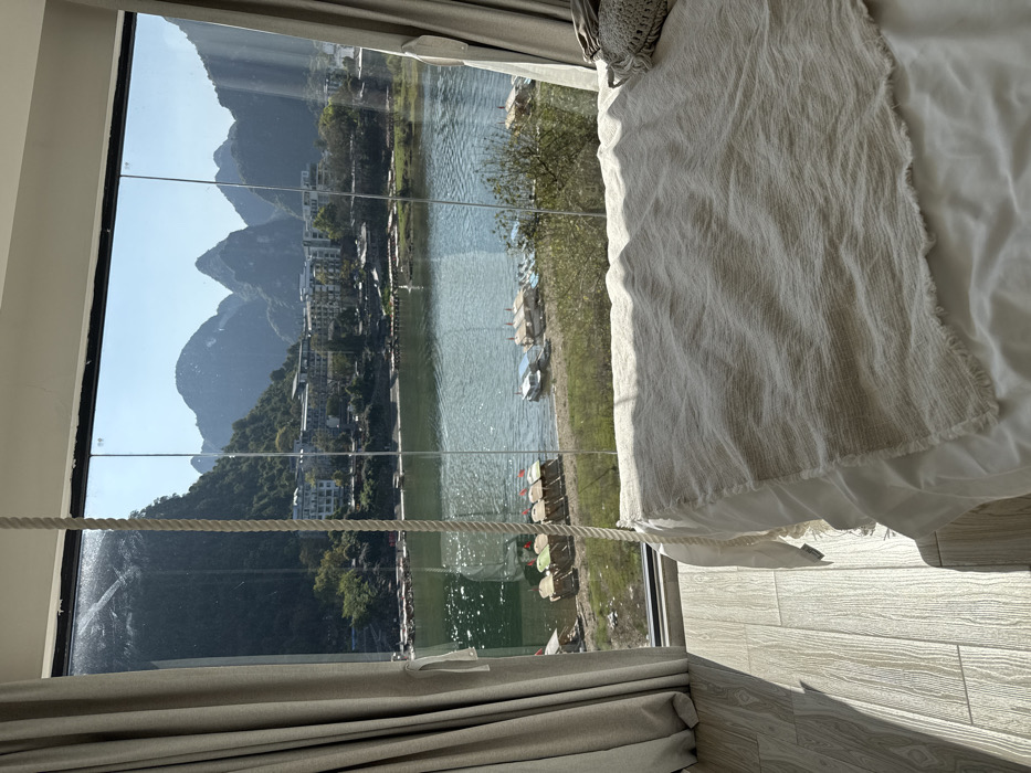 The image displays an interior room, likely a hotel bedroom, featuring a panoramic view of a river and mountainous landscape in Guilin, China. In the immediate foreground, a bed with white sheets and a textured, off-white throw blanket is positioned on the right. A thick, braided white rope hangs vertically from the ceiling in the center of the frame. The floor on the left consists of light-toned, wood-patterned tiles. Light-colored curtains are drawn back to frame a large, multi-pane glass window that extends from floor to ceiling.
Through the window, a wide body of water, identifiable as part of the Li River system, is visible across the midground. Several boats are moored along the riverbank; a cluster of smaller, flat-bottomed boats with distinct red, green, yellow, and white canopies is visible on the left, while larger, flat-decked vessels are situated further right. Beyond the river, multi-story buildings with light facades are constructed along the verdant riverbanks and at the base of prominent, green-clad hills. The background is dominated by the iconic, sharply rising karst limestone mountains characteristic of the Guilin region, silhouetted against a clear, bright blue sky. The scene is illuminated by strong natural daylight. No individuals are present in the image.