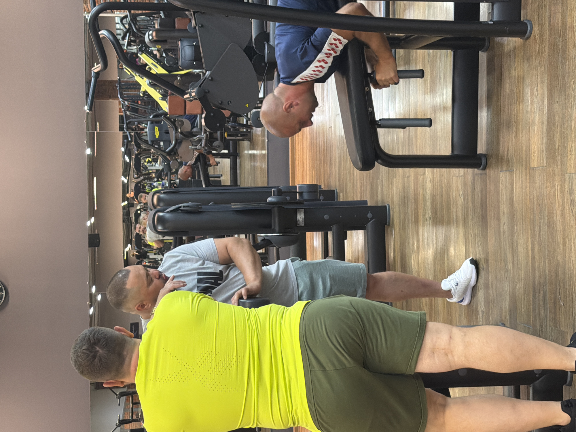 A group of four men are in a gym in Chaikovsky, Russia. Two men in the foreground are talking. One man is wearing a bright yellow t-shirt and olive green shorts, and the other is wearing a grey t-shirt and grey shorts. A third man is in the background, bent over a weight machine, wearing a blue and white t-shirt and black shorts. The fourth man is also in the background, using a weight machine. The gym has wooden floors and mirrors on the walls.