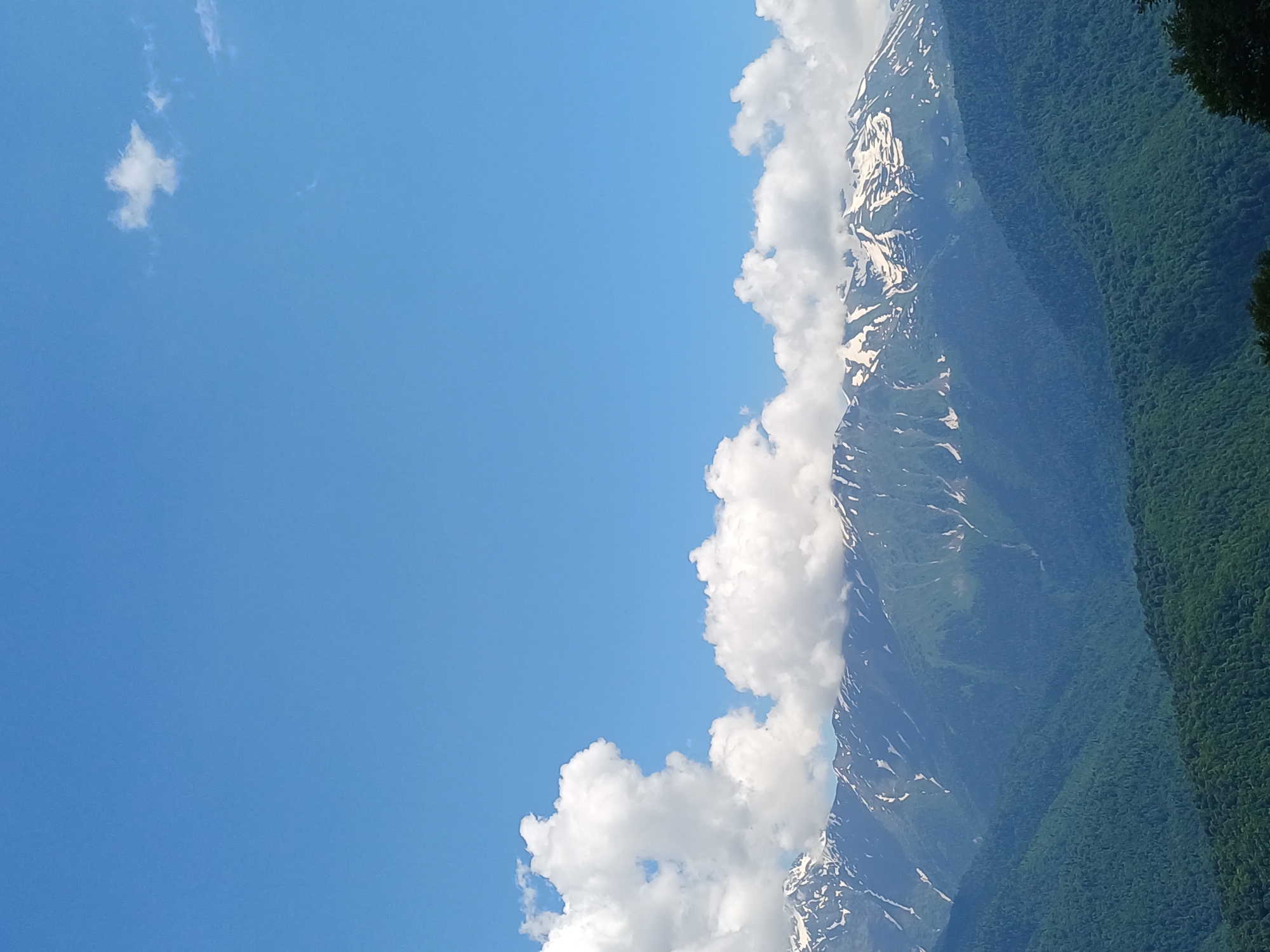 This image captures a majestic mountain landscape under a clear blue sky. The foreground features rolling green hills covered in dense forest. In the mid-ground, a range of mountains rises, their upper slopes dotted with patches of snow, indicating a high altitude. The peaks are rugged and dramatic. Large, fluffy white clouds are scattered across the sky, some obscuring parts of the mountain peaks. The overall scene suggests a serene and natural environment, likely a popular scenic viewpoint in the region of Estosadok, Russia, known for its mountainous terrain and proximity to ski resorts. No people or specific man-made objects are visible, emphasizing the natural beauty of the location.