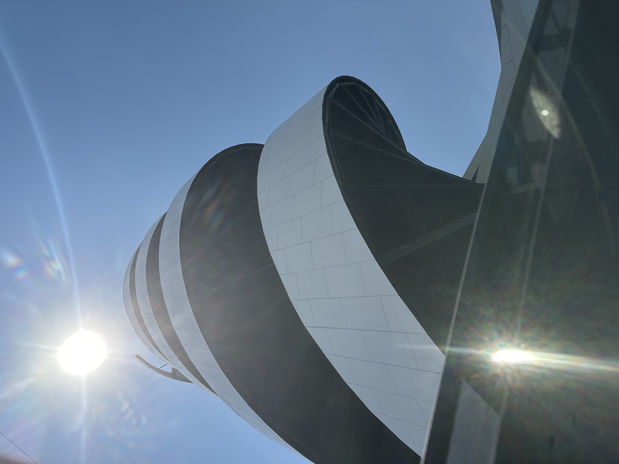This image captures a modern architectural structure in San Bernardino Tlaxcalancingo, Mexico, viewed from a low angle looking upwards against a clear, bright blue sky. The primary subject is a building featuring a distinctive, curved, and possibly spiraling design. Its facade consists of alternating bands of light-colored, possibly white or light grey, rectangular panels and darker, recessed or shadowed sections, creating a dynamic, layered effect.
The sun is prominently positioned in the left portion of the frame, appearing as a brilliant, starburst-like light source with significant lens flare and rays extending across the sky. This indicates bright, sunny weather, likely mid-day or early afternoon. The sky is entirely clear, devoid of clouds. The strong sunlight emphasizes the building's contours and textures.
No people or specific activities are visible, placing the focus entirely on the building's unique design and the natural light. The architecture suggests a modern, possibly iconic public building such as a museum or cultural center. No visible text is present in the image.