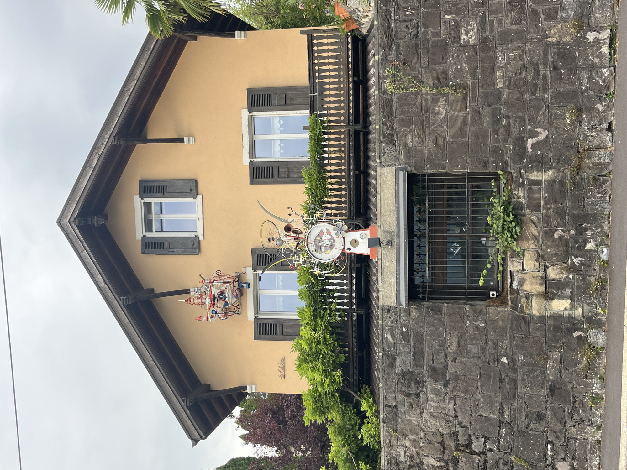 The image shows the facade of a building in Montreux, Switzerland, with a prominent artistic clock and sculptures. The building has a light orange stucco exterior, a dark wooden roof with exposed beams, and dark wooden shutters on the windows. A balcony with a decorative wooden railing runs across the upper floor, adorned with lush green plants. Above the balcony, on the left side of the building, is a red and white sculpture with various mechanical parts and figures. To the right of the balcony, a large, colorful clock with a face featuring cartoon-like characters and gears is mounted. The clock is integrated with bicycle wheels and other metalwork, creating an eccentric, whimsical art piece.
The building is partially built into a stone retaining wall, which forms the base of the scene. This wall is constructed from rough-hewn grey stones of varying sizes and shows signs of weathering and plant growth. A window with a black metal grate is set into the stone wall. The ground in front of the wall is paved, with a sidewalk visible. Trees and foliage, including a palm frond, frame the building and the stone wall, suggesting a temperate climate. The sky is overcast, with a grey, cloudy appearance. The overall impression is of a residential building with unique, artistic embellishments, set against a rugged, naturalistic backdrop. The sign "La Colette" is visible on the left side of the building's facade.