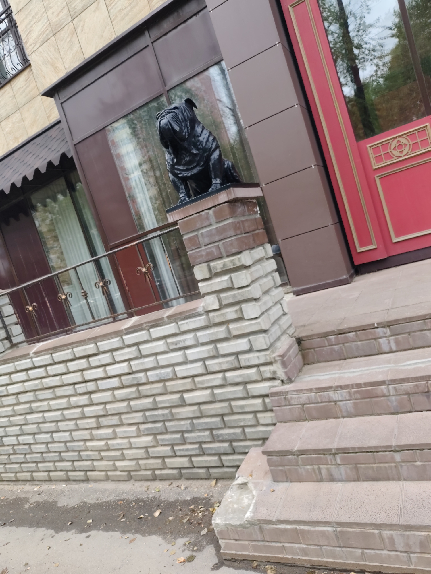 The image depicts the exterior of a building with a prominent statue of a black bulldog. The building features a facade of beige and brown panels, with large windows and a red door adorned with gold trim. A decorative metal railing is visible on a balcony or elevated walkway. The ground level is clad in light-colored brickwork, and there are steps leading up to the entrance. The environment appears to be urban, with some trees reflected in the windows. The specific location in Syktyvkar, Russia, is not determinable from the image alone. No people are visible in the scene.