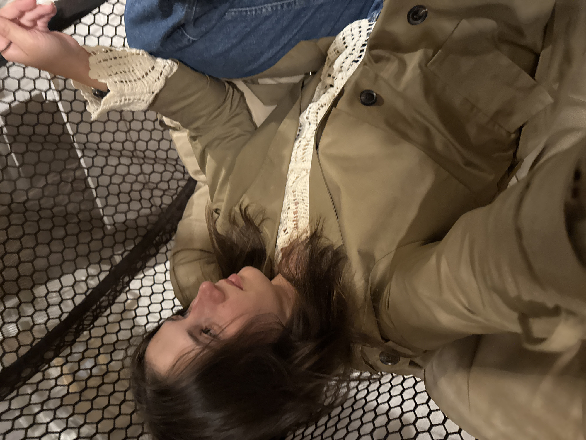 The image captures a young woman lying down on her side, positioned diagonally across the frame. Her head is towards the lower right, with her face in profile looking slightly upwards. She has dark brown hair and a calm, relaxed expression.
She is dressed in a khaki or olive green jacket or coat, with a cream-colored, possibly crocheted, top or blouse visible underneath. Blue denim jeans are also visible on her lower body. Her left arm is slightly raised, revealing the intricate crochet detail on the cuff of her top.
The woman is resting on a surface characterized by a distinct black hexagonal mesh pattern, which covers much of the background and foreground. This mesh suggests she might be on a modern bed frame, a hammock-like structure, or a piece of furniture with a hexagonal design. The lighting in the image is dim, indicating an indoor setting or possibly evening hours. There are no visible location cues within the image itself to confirm the provided context of Perm, Russia, and no discernible text. The overall atmosphere is one of casual relaxation.