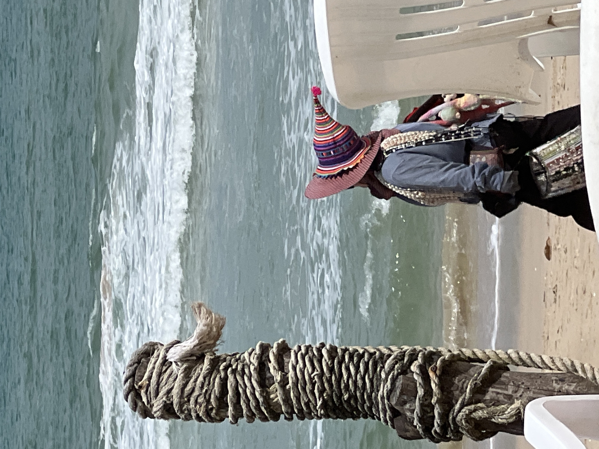 A person walks on a beach near the ocean. The person wears a blue shirt, dark pants, and a colorful, conical hat. They are carrying multiple bags. In the foreground, a wooden post wrapped with thick rope stands to the left. To the right, a white plastic chair is partially visible. The ocean has waves and the sand is light brown. The sky is not visible in the frame.