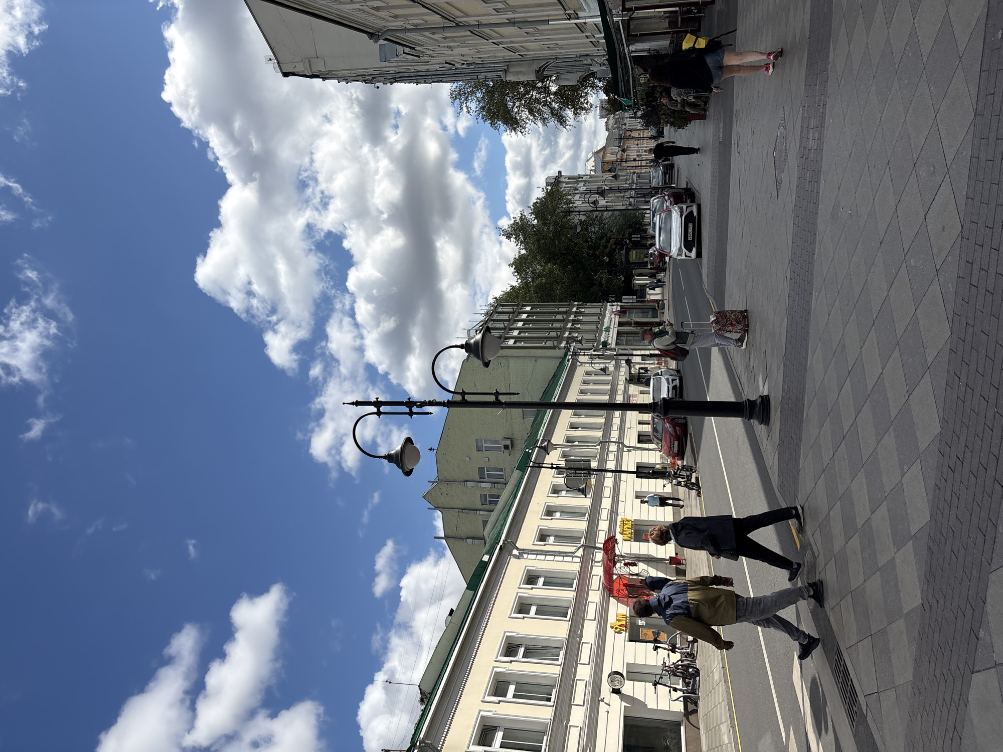 The image captures a street scene in Moscow, Russia, under a partly cloudy blue sky. The perspective is from a sidewalk looking down a moderately wide street lined with buildings of varying architectural styles and heights. The buildings on the left are older, pale yellow structures with multiple windows and green roofs. A storefront with a red awning is visible. On the right, the buildings are taller and appear more modern, with some balconies.
The street itself is paved with asphalt, and there is a pedestrian crosswalk in the foreground. Two people are walking across the crosswalk from left to right. The person on the left is wearing a brown and blue jacket and grey pants, while the person on the right is wearing a dark jacket and black pants. Both are walking briskly.
Further down the street, several cars are parked or driving. A white sedan is in the foreground on the right side of the street. Several lampposts with traditional designs are spaced along the sidewalk. Trees with green foliage are visible in the distance, interspersed between buildings. The sidewalk is paved with large gray tiles and some brick accents.
A woman is walking on the right sidewalk further down the street, carrying a yellow bag. Another person is walking away from the camera on the right side of the street. Several tables and chairs are set up outside a building on the right, suggesting an outdoor seating area for a cafe or restaurant. The overall atmosphere is that of a bustling urban environment on a pleasant day.
