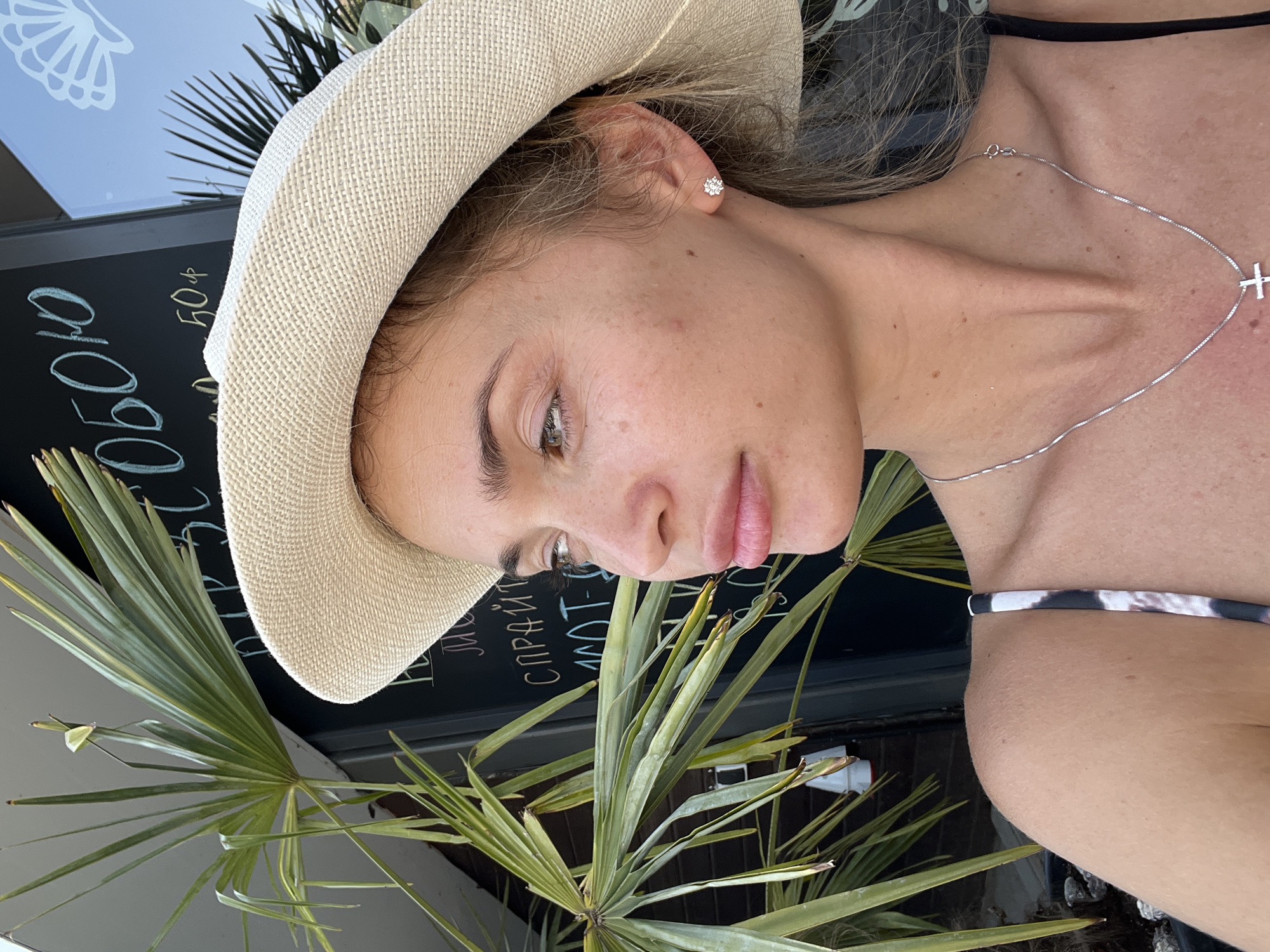A woman is posing for a selfie, looking to her right. She is wearing a wide-brimmed straw hat and a silver necklace with a cross pendant. The background shows a blackboard with text written on it, some palm fronds, and a glimpse of a building. The lighting suggests it is daytime. The scene appears to be outdoors, possibly in a cafe or shop. The woman's location is identified as Fontanka, Ukraine.