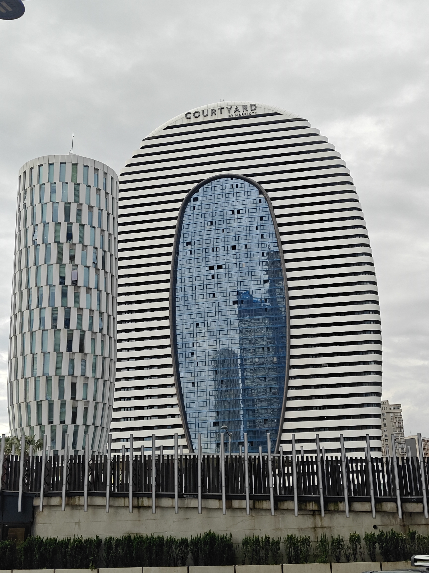 The image captures a view of two distinct modern buildings under an overcast, light gray sky. In the foreground, a dark metal barrier or fence with vertical posts, some of which are light silver-colored, is elevated on a concrete structure. Below this concrete wall, a line of green hedges or bushes is visible.
The building on the left is a tall, cylindrical structure with a facade characterized by alternating vertical white panels and narrow, light blue or gray window sections. This creates a rhythmic, patterned appearance.
The building on the right is the dominant feature, showcasing a highly distinctive and curved architectural design. It has a broad, almost oval-like profile, with its exterior covered in prominent horizontal white bands. A large, central section of this building features a dark blue, oval-shaped glass facade. This glass surface reflects parts of the cloudy sky and other structures, including what appears to be another building with a blue protective covering or a blue facade.
Above the large glass section on the right building, the word "COURTYARD" is clearly visible in a dark, capital letter font, with "BY MARRIOTT" printed in smaller text directly below it, identifying the building as a Courtyard by Marriott hotel. The absence of direct sunlight and the uniform cloud cover suggest a cloudy daytime setting. No human activity or specific events are depicted, placing the focus entirely on the architectural subjects and the urban environment.
