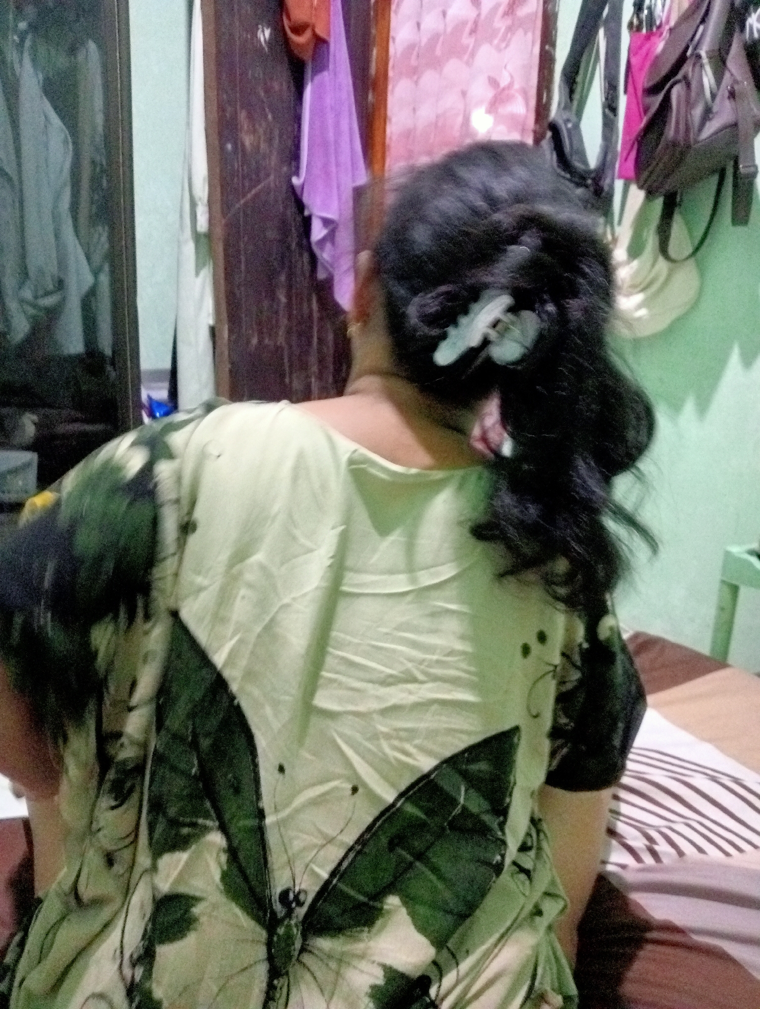 A woman with dark, wavy hair adorned with a light-colored butterfly clip is seen from the back, wearing a light green blouse with a repeating pattern of dark green butterflies. She is sitting on a bed with a striped comforter. The background appears to be a room with a wooden closet, and clothing hanging on hangers. A portion of what might be a mirror or a closet door is visible to the left. The overall impression is an indoor setting in a residential area. The location provided is Kota Negara Timur, Indonesia, suggesting this scene is within that geographical area.