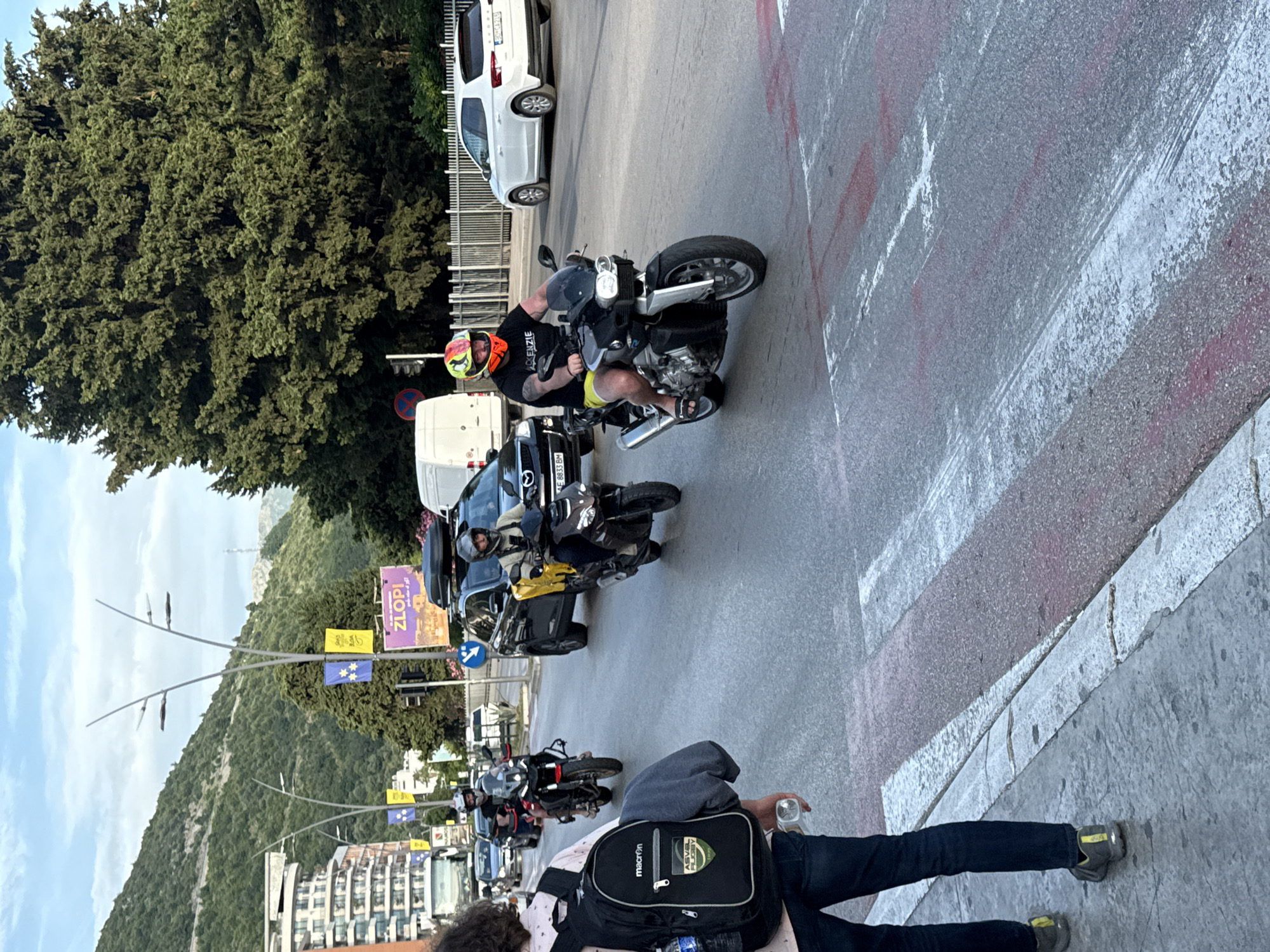 The image shows a street scene in Budva, Montenegro. Several people on motorcycles are navigating through traffic. In the foreground, a person with a backpack is walking on the sidewalk. In the background, there are buildings, trees, and a mountain. The weather appears to be partly cloudy.