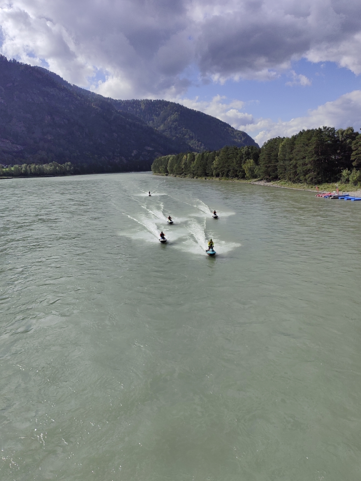 An aerial view captures a wide, choppy river flowing through a mountainous landscape. The river's water is a murky, light green color, with visible ripples and spray from several jet skis. Five individuals are riding jet skis, each creating a white wake behind them. The jet skis are spaced out along the river, moving in the same direction. On the right bank of the river, a dense forest of green trees lines the shore. In the distance, a steep, dark green mountain rises, partially obscured by a dramatic sky filled with white and dark gray clouds. Several small, colorful floating objects, possibly buoys or personal watercraft, are visible near the shore on the right side. The overall scene depicts a recreational activity on a natural waterway.