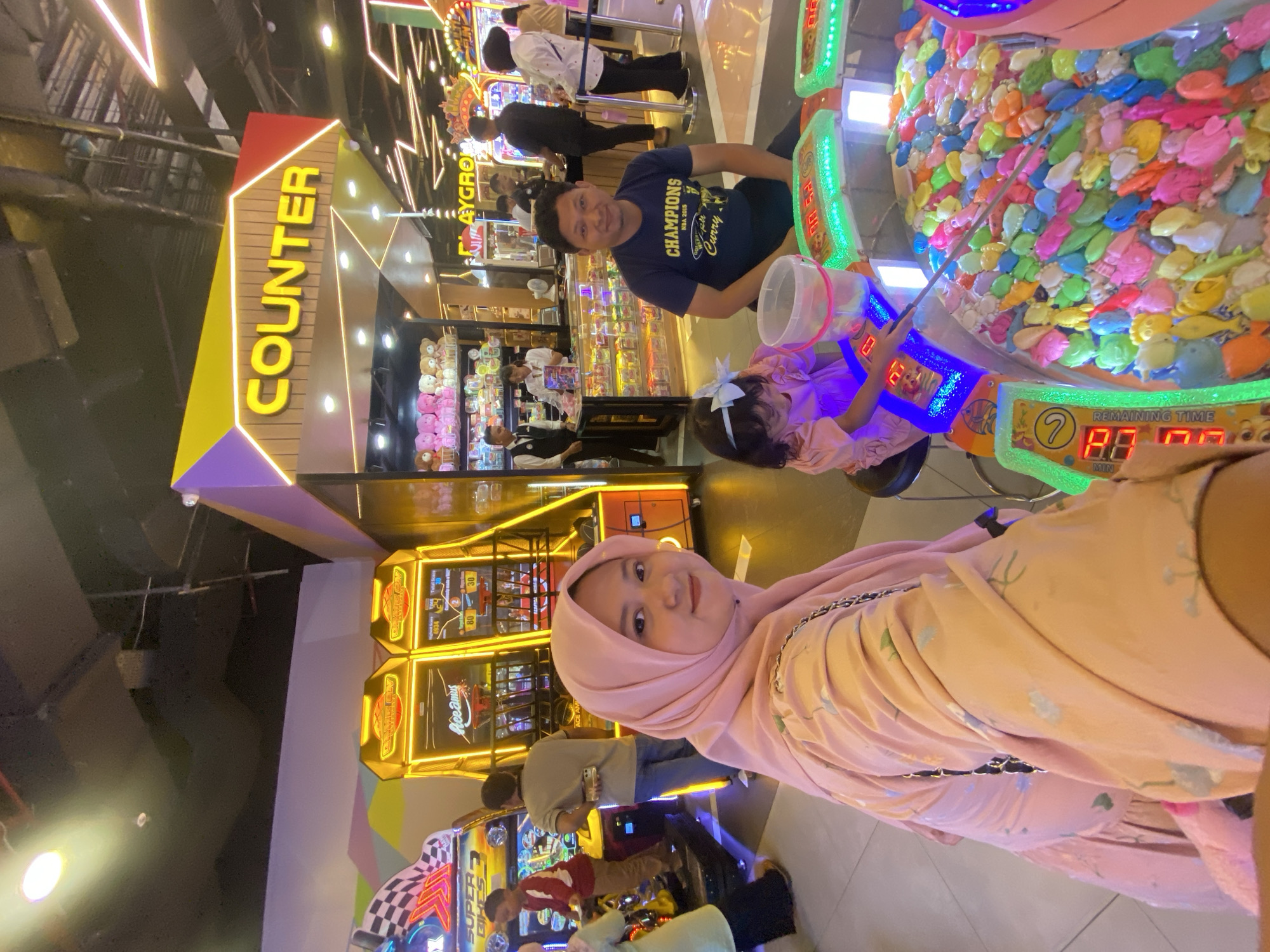 The image depicts an indoor entertainment arcade in Sidoarjo, Indonesia. In the foreground, a woman wearing a pink hijab and dress is looking towards the camera, with her arm outstretched. To her right, a man in a blue t-shirt is seated, facing forward. In front of them, a young girl in a pink dress is playing a fishing game, holding a fishing rod over a pool filled with colorful plastic sea creatures. Behind the family, several arcade game machines are visible, including a racing game and a basketball game. Further in the background, a counter area displays shelves stocked with plush toys and other merchandise. The environment is brightly lit with neon signage and fluorescent lights, characteristic of an arcade.