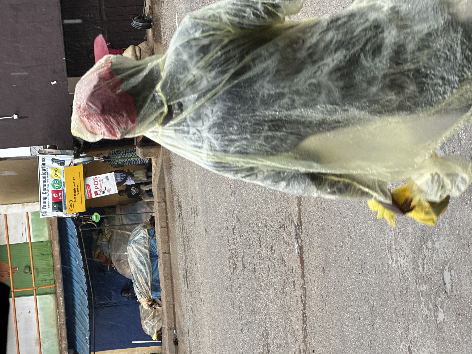 In Bukuru, Nigeria, a person wearing a yellow disposable raincoat walks away from the camera on a wet asphalt road. They are wearing a red baseball cap under the hood of the raincoat. In their right hand, they are carrying a bunch of yellow items, possibly flowers or produce. Behind the person, a blue awning covers a storefront with various items, including large bags covered in plastic sheeting, piled up. To the left of the awning, a green metal gate with orange trim is visible. A sign for "DC Young Communications SIM REGISTRATION AND POS SERVICES" with logos for Airtel, Glo, and MTN is attached to the building. Another sign below it advertises "MTN SIM Registration Center." Further down the street, a person is seated under the awning next to some of the covered items, and another person is visible further back in the shop. The ground is damp, indicating recent rain, and the weather appears overcast.