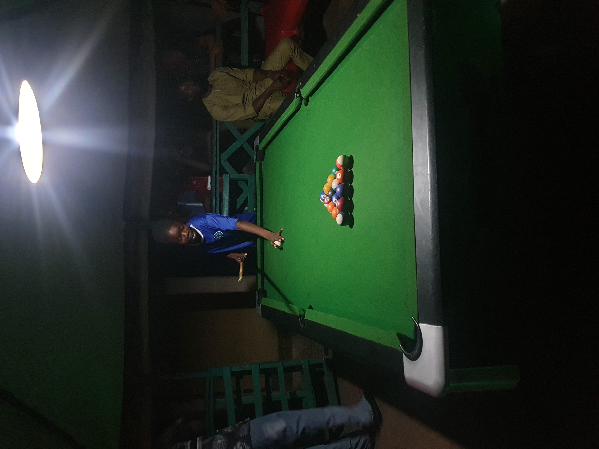 A young Black person in a blue shirt is playing billiards indoors. They are positioned at the far end of the table, about to strike a white cue ball with a cue stick. A rack of colored billiard balls is set up near the center of the green felt surface. In the background, other individuals are visible, including a Black person in a light brown tunic sitting on a bench and another person partially visible on the left side of the frame. The environment appears to be dimly lit with a bright overhead light illuminating the billiard table. The scene suggests a casual gathering or recreational activity taking place in Lokoja, Nigeria.