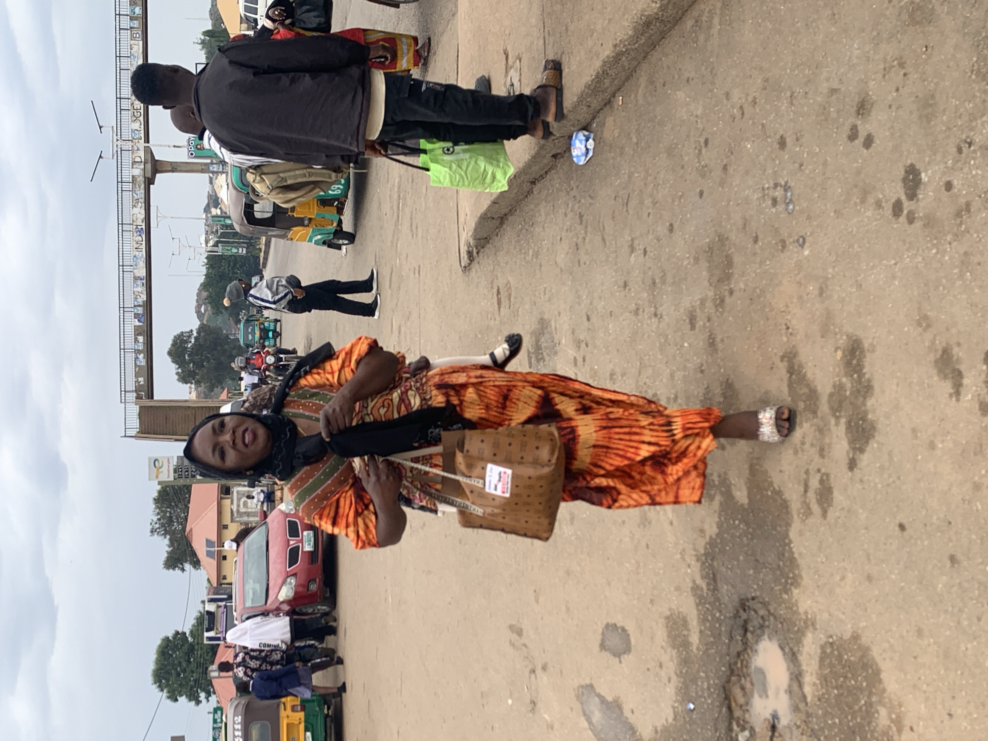 The image shows a street scene in Gura Top, Nigeria. A woman in an orange and yellow patterned dress and a black headscarf is walking towards the camera, carrying a brown patterned handbag. She is wearing jeweled sandals. In the background, there are various vehicles, including a red car, a yellow and green tricycle, and motorcycles. Several people are visible walking and standing on the street and sidewalk. An elevated walkway or bridge is visible in the background. The ground appears to be paved with asphalt and has some puddles and stains. The sky is overcast.