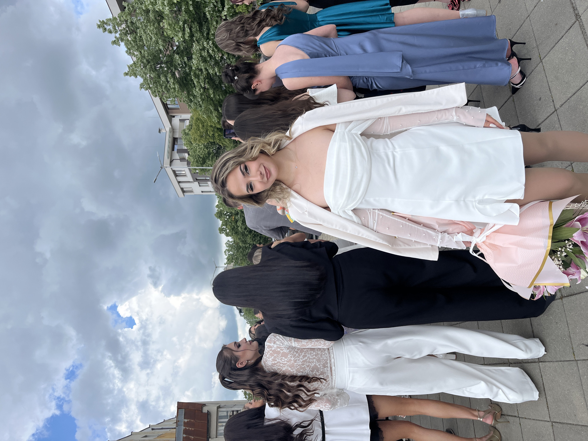A group of young women are standing on a paved surface, likely a sidewalk, in Blagoevgrad, Bulgaria. Buildings and trees are in the background, with a cloudy sky overhead. They are posing for a photo; some are holding bouquets of flowers. The woman in the front is smiling at the camera, wearing a white dress with a light pink sash, and a white jacket. The women behind her are wearing formal dresses in shades of blue, black, and white.