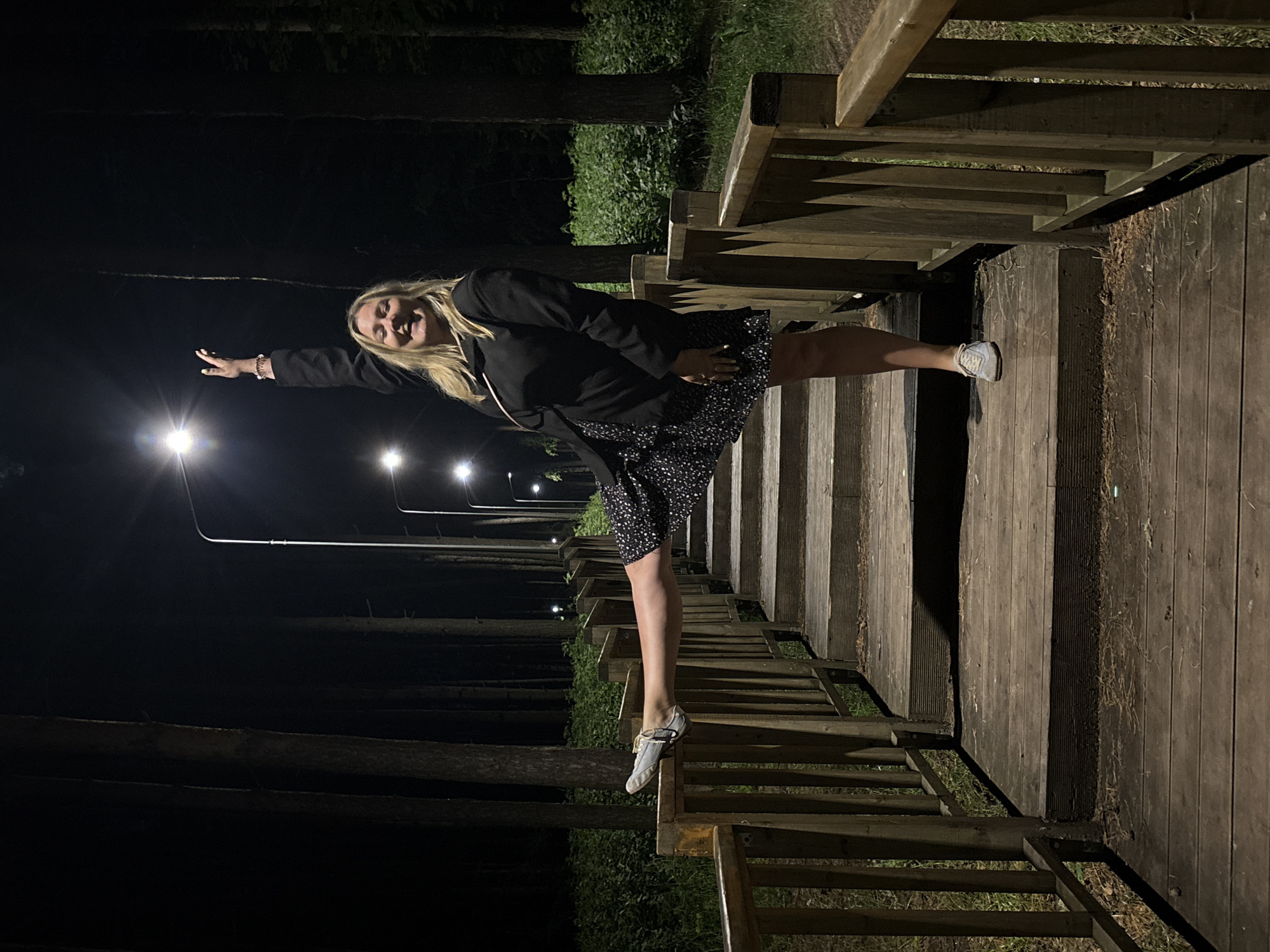 An adult female is standing on a wooden staircase in a park at night. She is wearing a black jacket and a black and white polka dot dress. Her right leg is extended and resting on a wooden railing. She is smiling and her left arm is raised. There are streetlights visible in the background, illuminating the path. The staircase is surrounded by trees. This image was captured in the city of Chaikovsky, Russia.