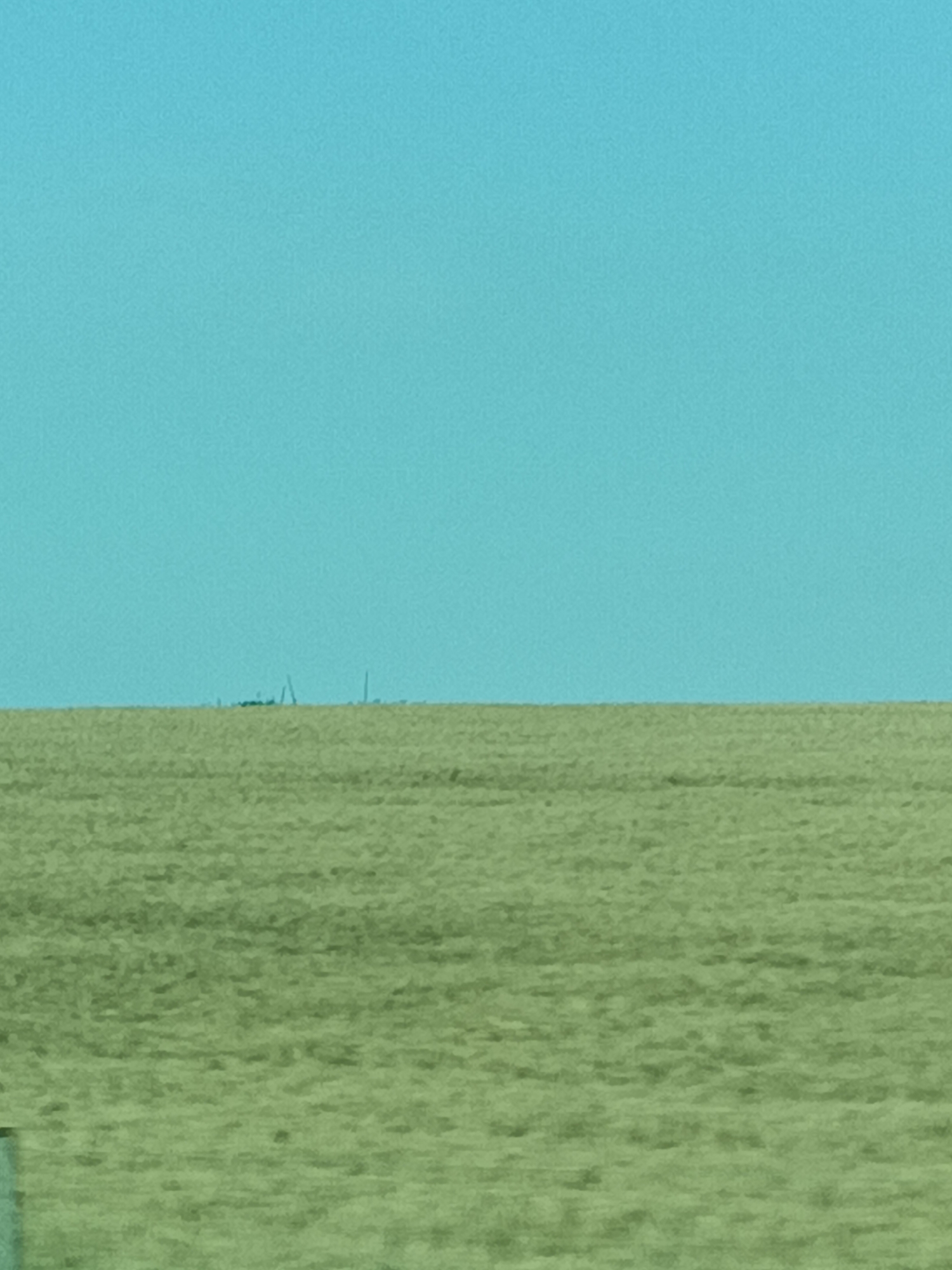 A distant, hazy line of industrial structures and possibly wind turbines is visible on the horizon, appearing as dark silhouettes against a bright, clear, turquoise sky. The foreground is dominated by a vast, flat field of dry, light green grass or crops, showing subtle undulations. The overall impression is one of a very wide-open, rural landscape under a bright, possibly hot, day. The low resolution and distance of the industrial elements suggest they are many kilometers away, possibly near Třemošnice, Czechia, and atmospheric conditions are creating a mirage-like effect. No people are visible, and the scene is devoid of any discernible activity or text.