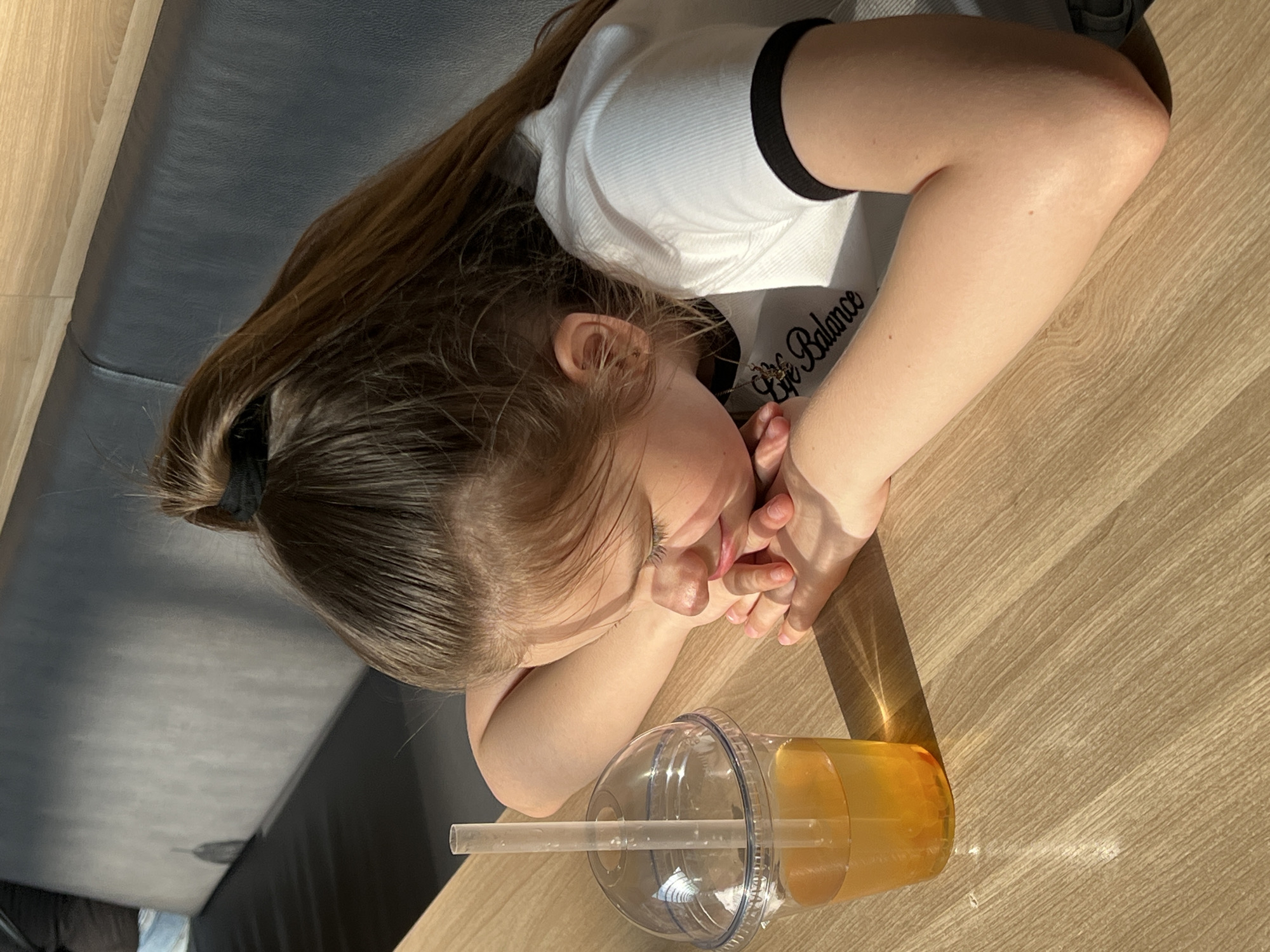 A young girl rests her head on a wooden table next to a plastic cup with a straw containing a yellow liquid. The girl has brown hair pulled back with a black hair tie and is wearing a white t-shirt with a black trim. She is resting her head on her hands. Part of a black upholstered seating is visible on the left side. The setting is likely a cafe in the city of Izhevsk, Russia.