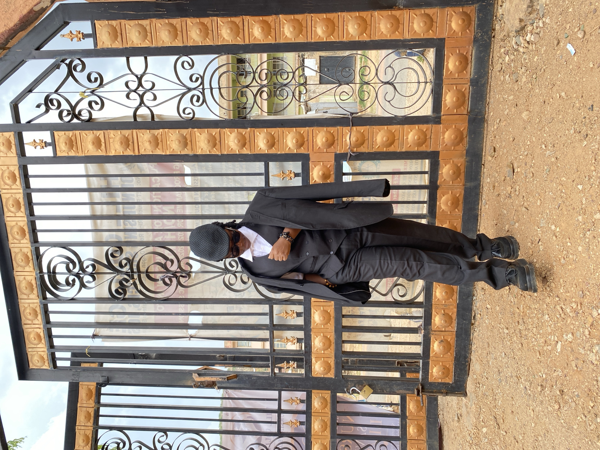 A man in a dark suit, white shirt, and hat stands in front of a large metal gate with decorative scrollwork. The gate is framed with orange square tiles. The location is Gwong, Nigeria. The ground is gravelly dirt. The man is wearing black shoes and bracelets. He has sunglasses on. His jacket is draped over his shoulders. A padlock secures the gate.