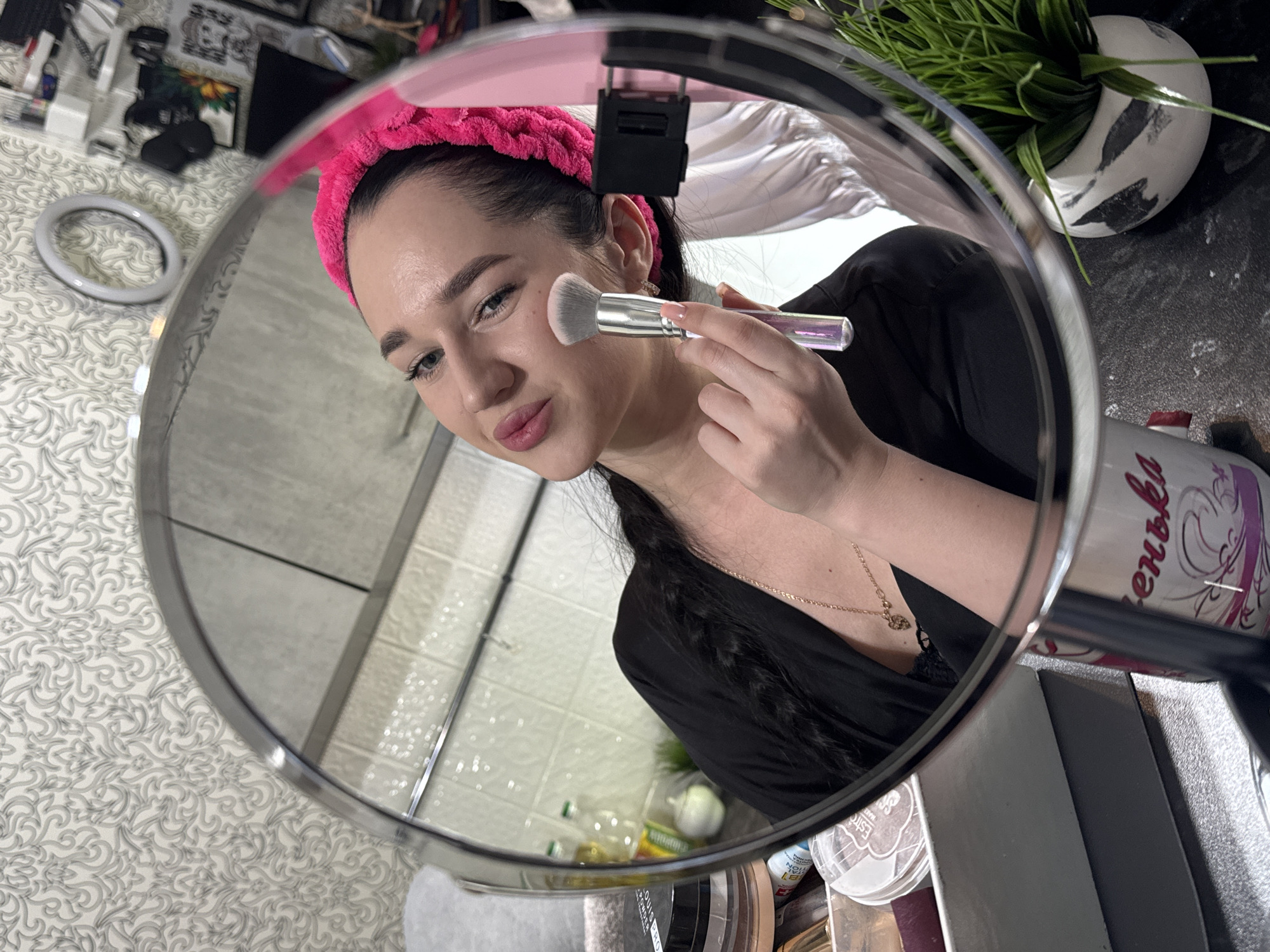 A young woman with a pink headband is applying makeup to her cheek with a brush, reflected in a large circular mirror. She is wearing a black robe and a gold necklace. The background reflected in the mirror shows a tiled wall, shelves with various items including bottles and makeup products, and a ring light. The foreground of the image shows a portion of the mirror stand and a mug with Cyrillic writing. The scene appears to be in a home environment, likely a bathroom or dressing area. There is no indication that the location is specifically in Novosibirsk, Russia.