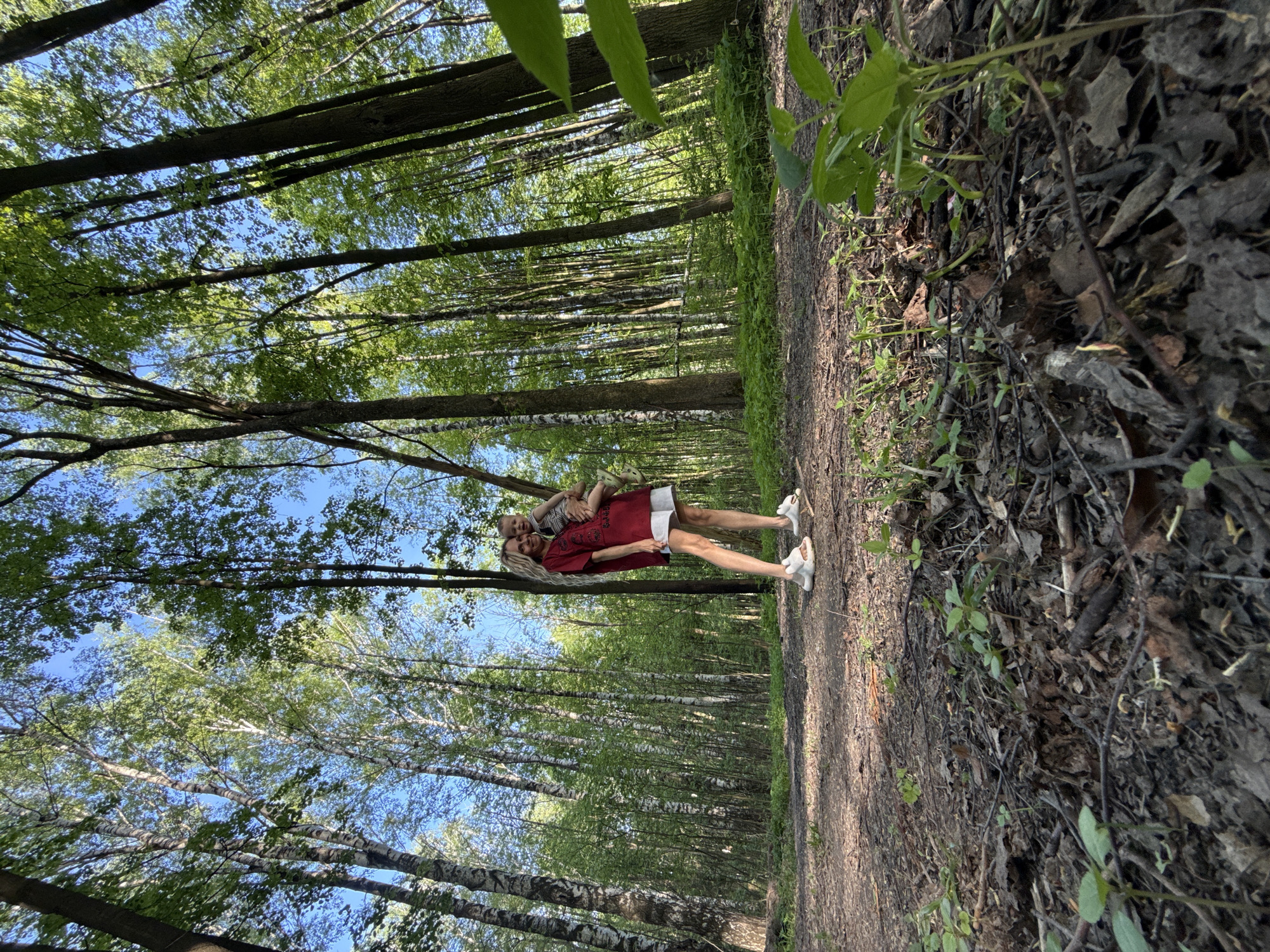A full body shot depicts a woman and a small child standing on a dirt path in a forest. The woman wears a red dress, white shorts, and white sneakers. The child wears a striped shirt and light-colored pants and shoes. The woman is facing the viewer with the child clinging to her back. The forest is dense with tall, thin trees and green leaves. The ground is covered in leaves and dirt. The sky is visible through the trees. The image suggests it was taken in Rusanovo, Russia based on location metadata.