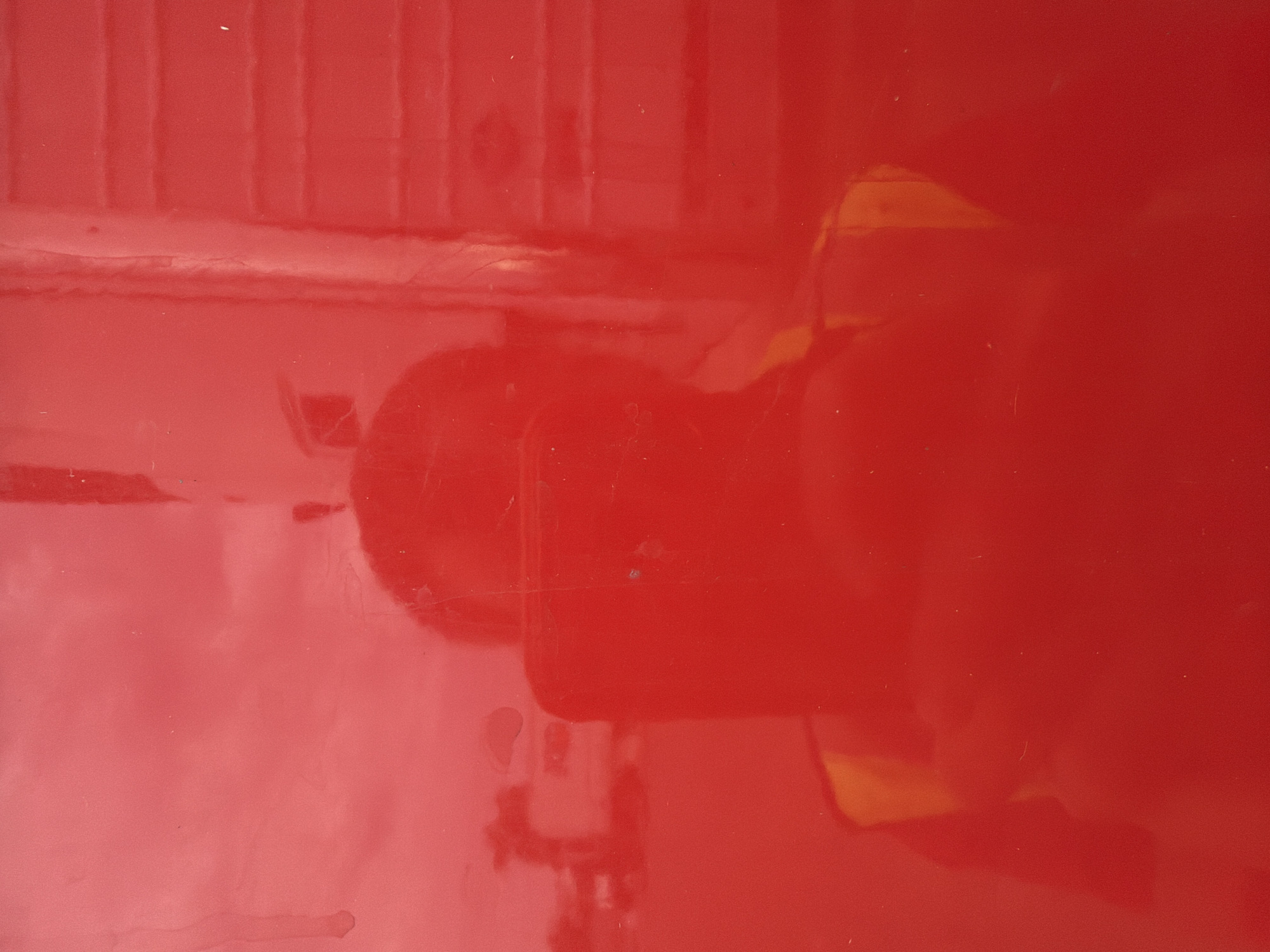 The image is a reflection on a red, glossy surface, possibly a vehicle. A person is visible in the reflection, holding a smartphone and wearing a safety vest. In the background, a building or structure with horizontal lines, possibly a trailer or part of a building, is reflected, along with some indistinct shapes that could be trees or other vehicles. The overall scene suggests an outdoor, possibly urban or industrial, setting in Murfreesboro, United States of America.